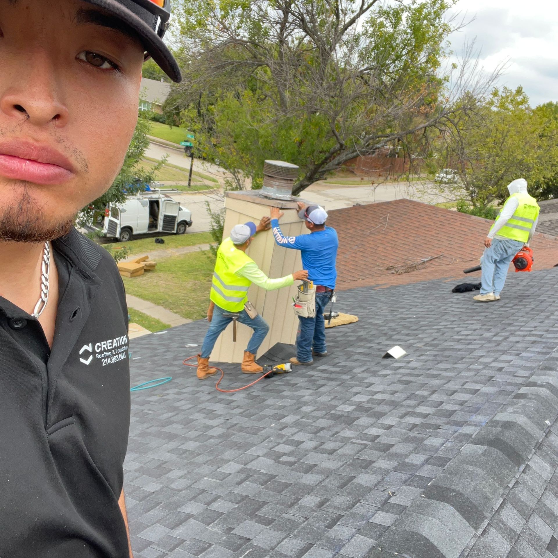 Man taking selfie on roof with construction workers repairing the roof. Green and blue work attire.