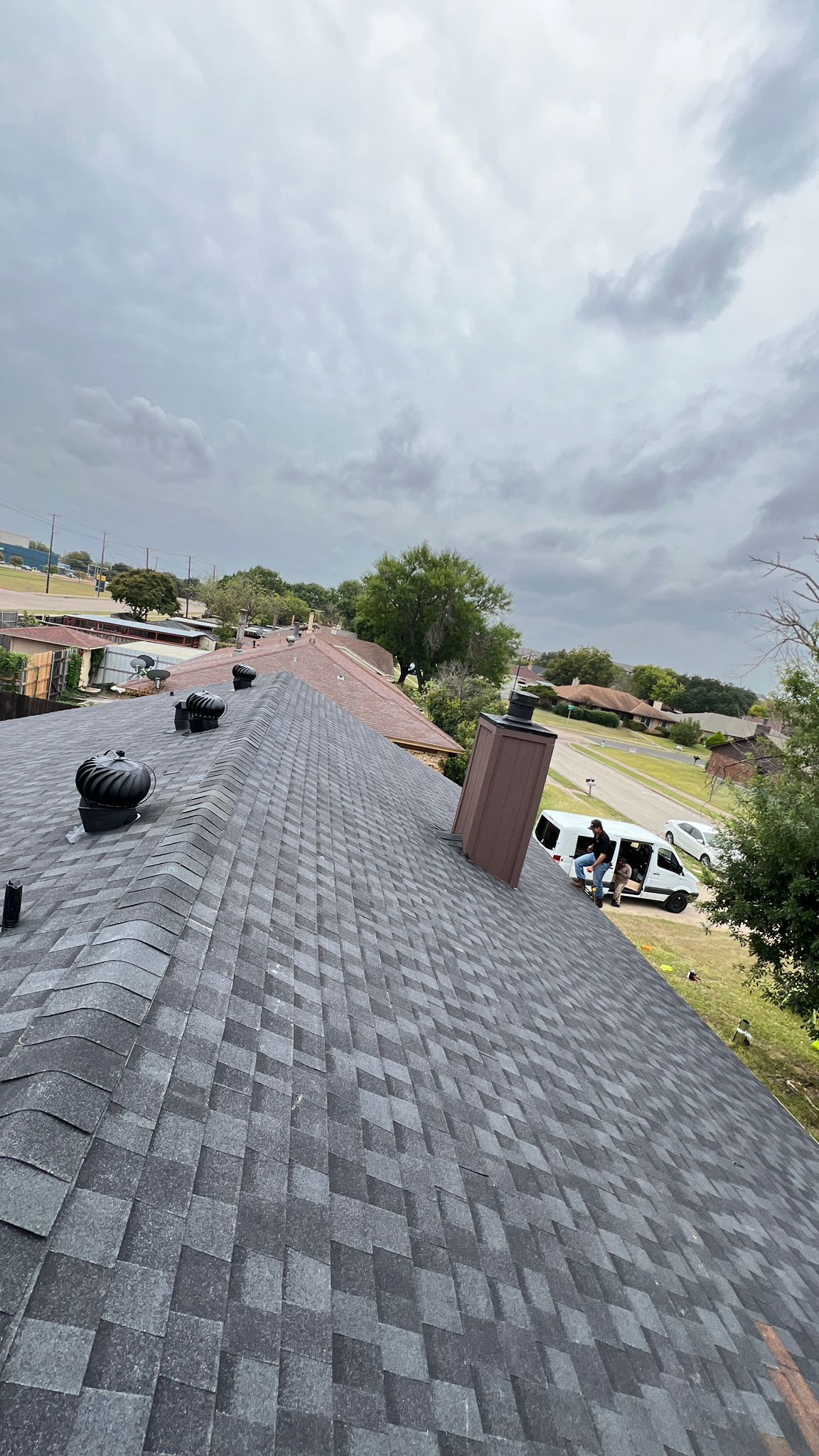 View of a roof with dark gray shingles, a chimney, and a cloudy sky.