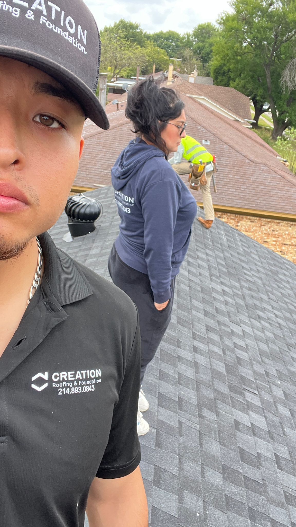 Two people on a rooftop inspecting shingles; man in cap and shirt looking at camera, woman in blue jacket.