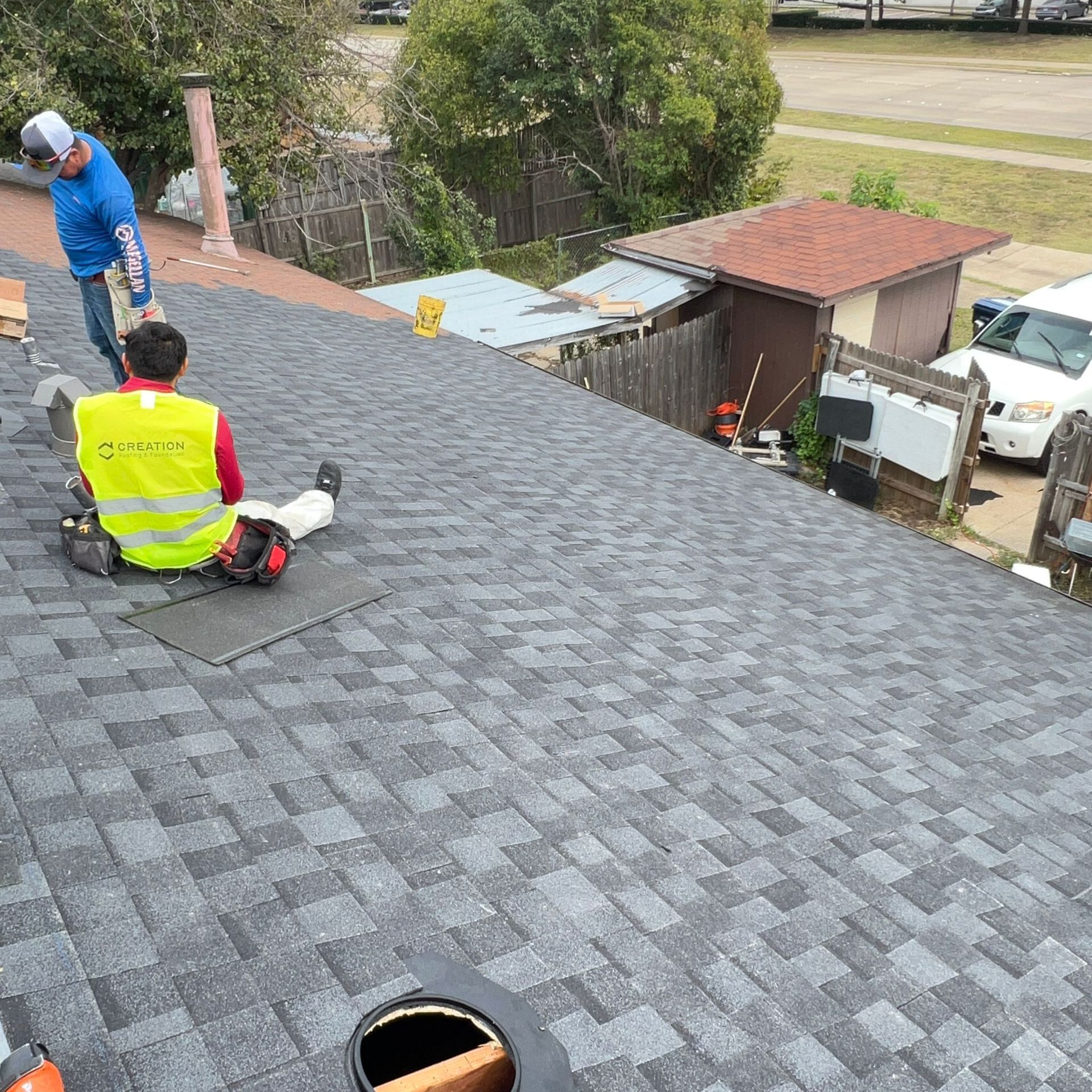 Two roofers on a newly shingled roof. One is holding something, the other is sitting. Overcast day.