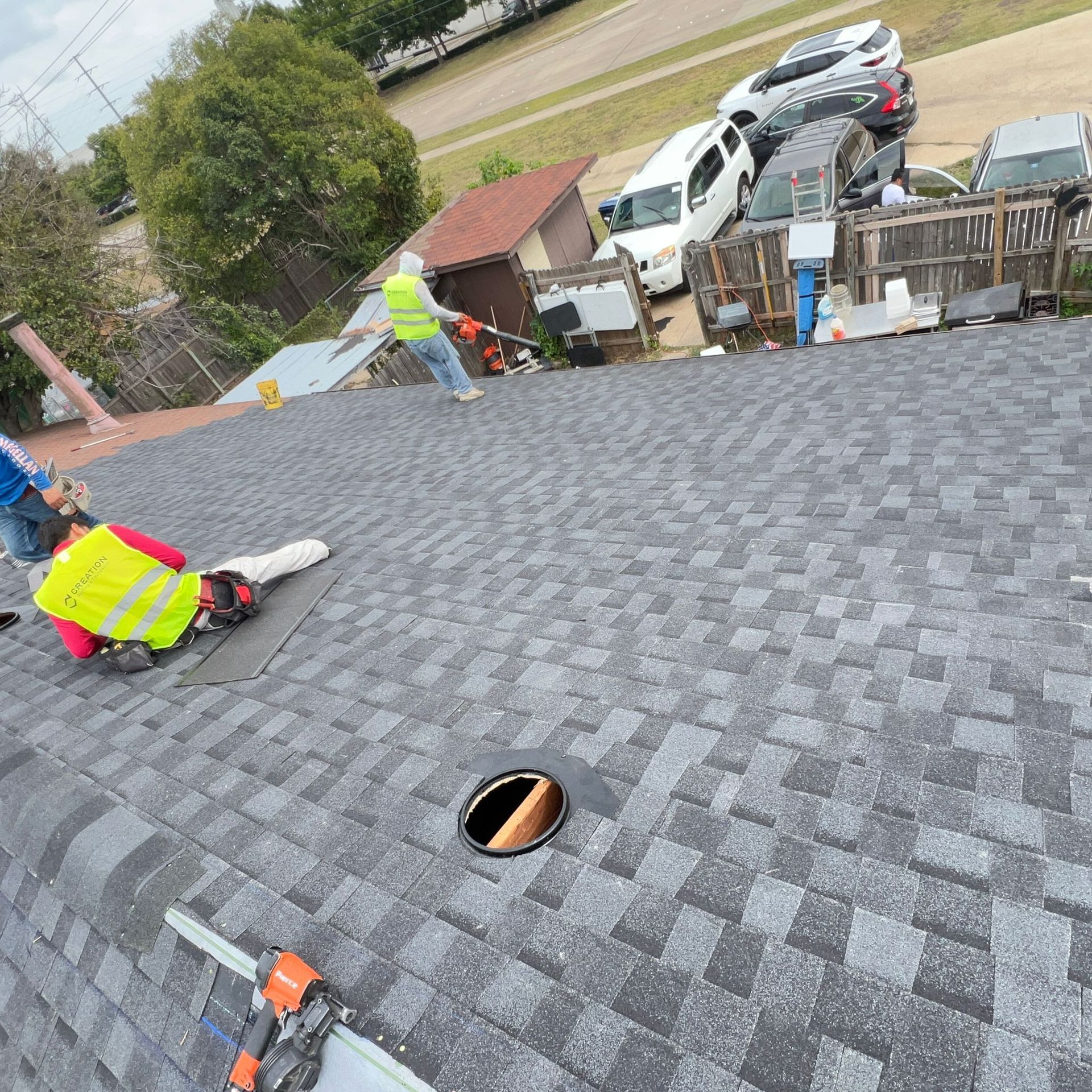 Workers in safety vests on a roof, installing gray asphalt shingles. Cars are parked nearby.