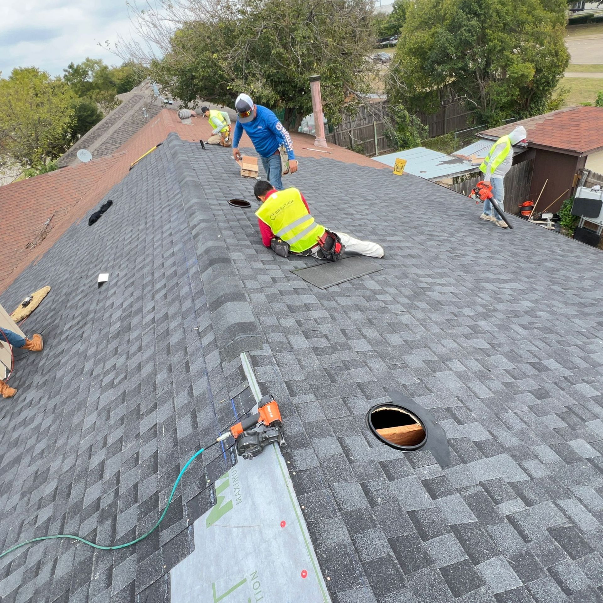 Roofers installing shingles on a house roof. Workers wearing safety vests, using tools, in a suburban setting.