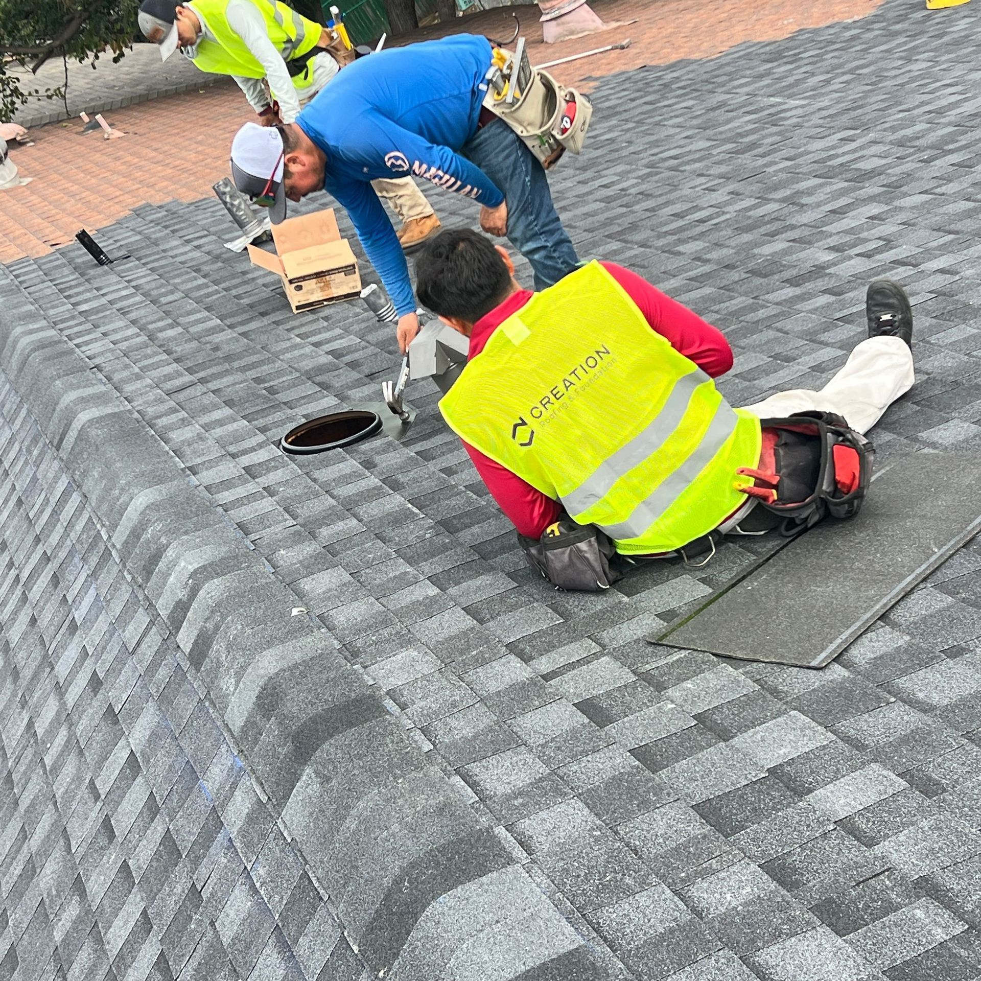 Roofers installing shingles on a house roof. One worker kneels, another stands, wearing safety vests.