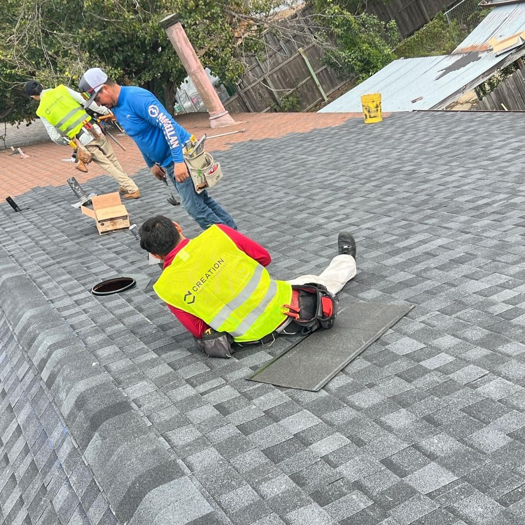 Roofers on a gray shingle roof, one kneeling, wearing safety vests, and another standing.