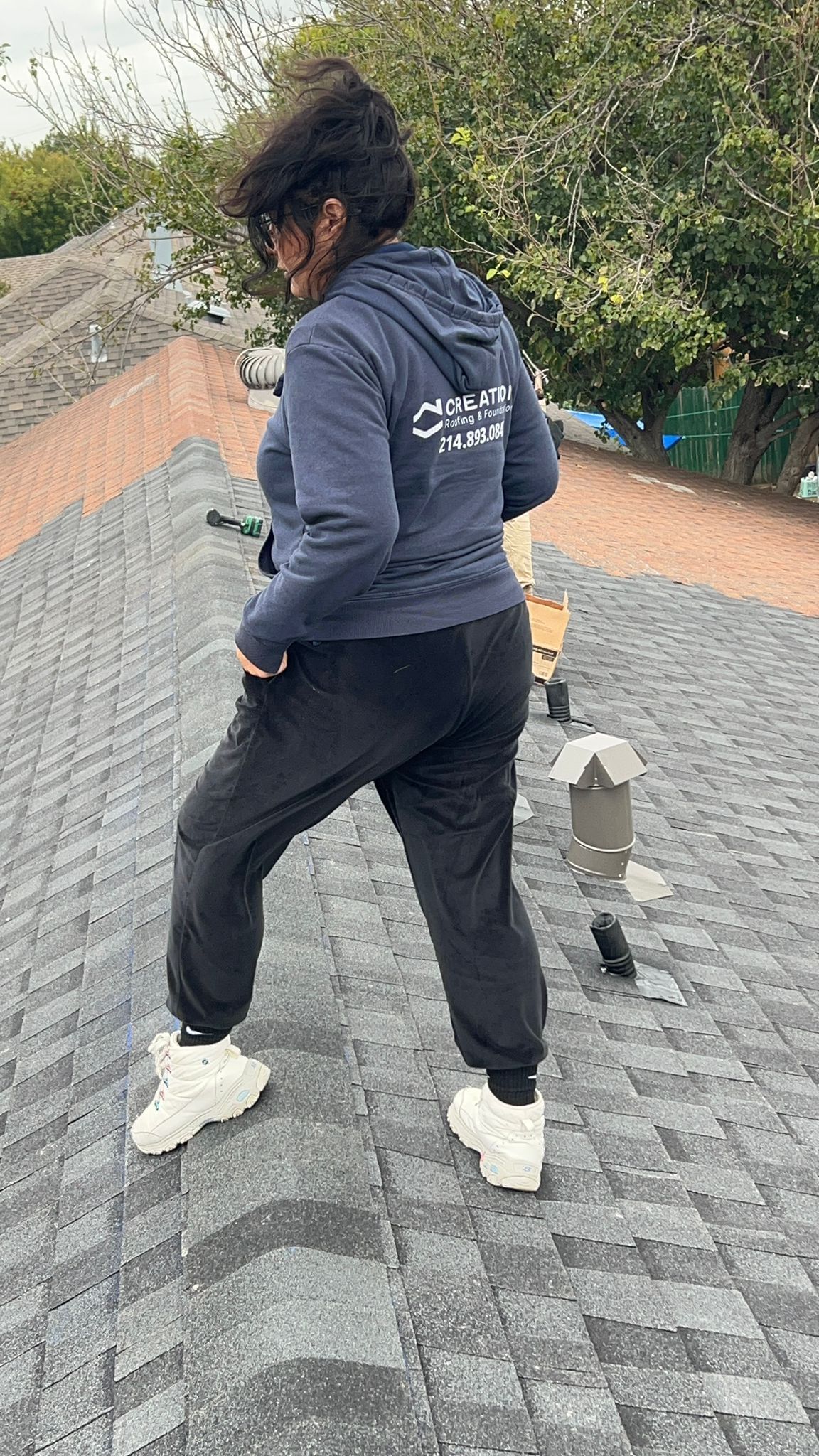 Person standing on a dark shingled roof, wearing a blue hoodie and black pants. White sneakers. Outdoor setting.