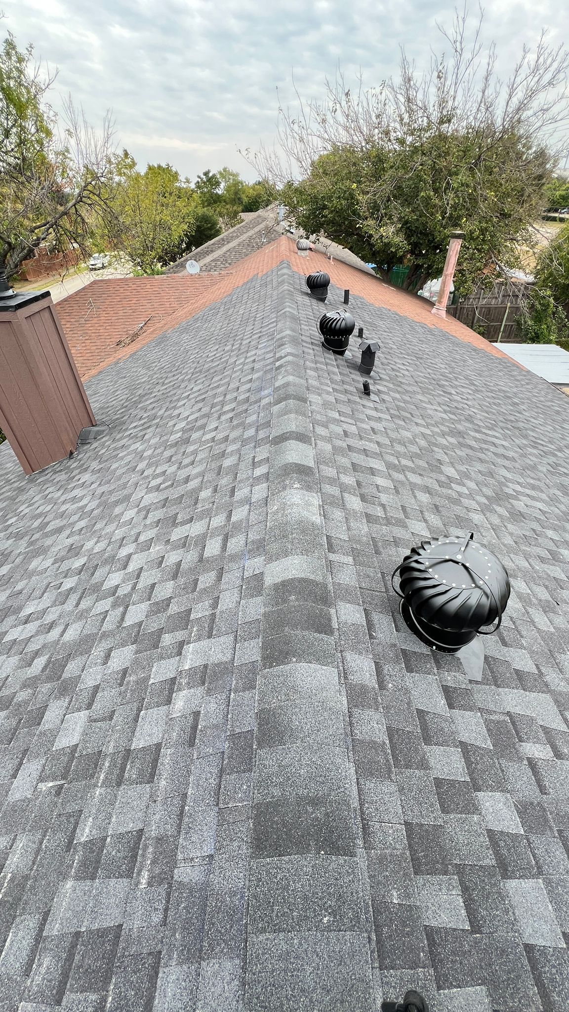Gray shingled roof with a brown chimney and several black vents, set against a cloudy sky and green trees.