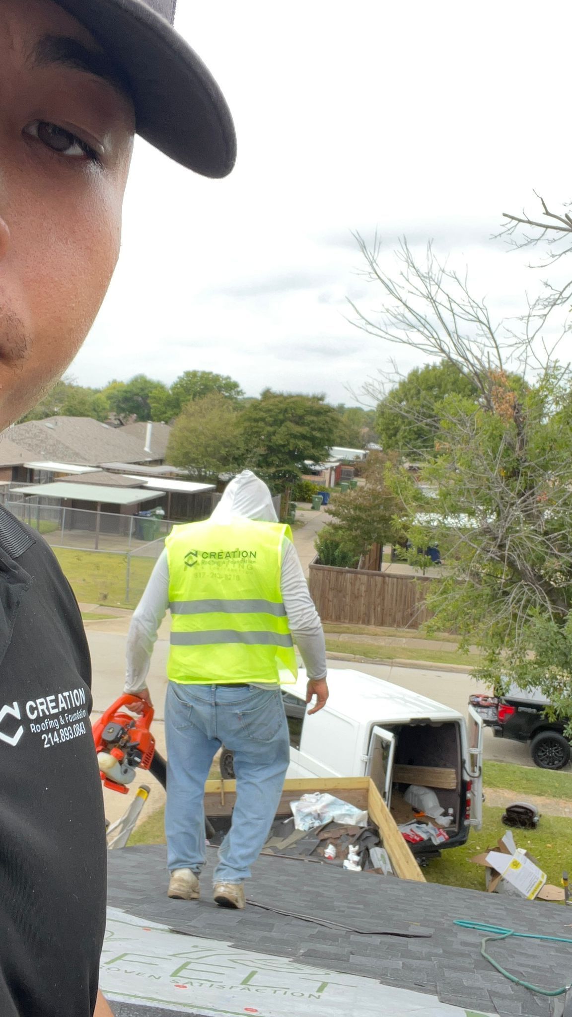 Man taking selfie on roof; worker in yellow vest carries blower toward van on rooftop.
