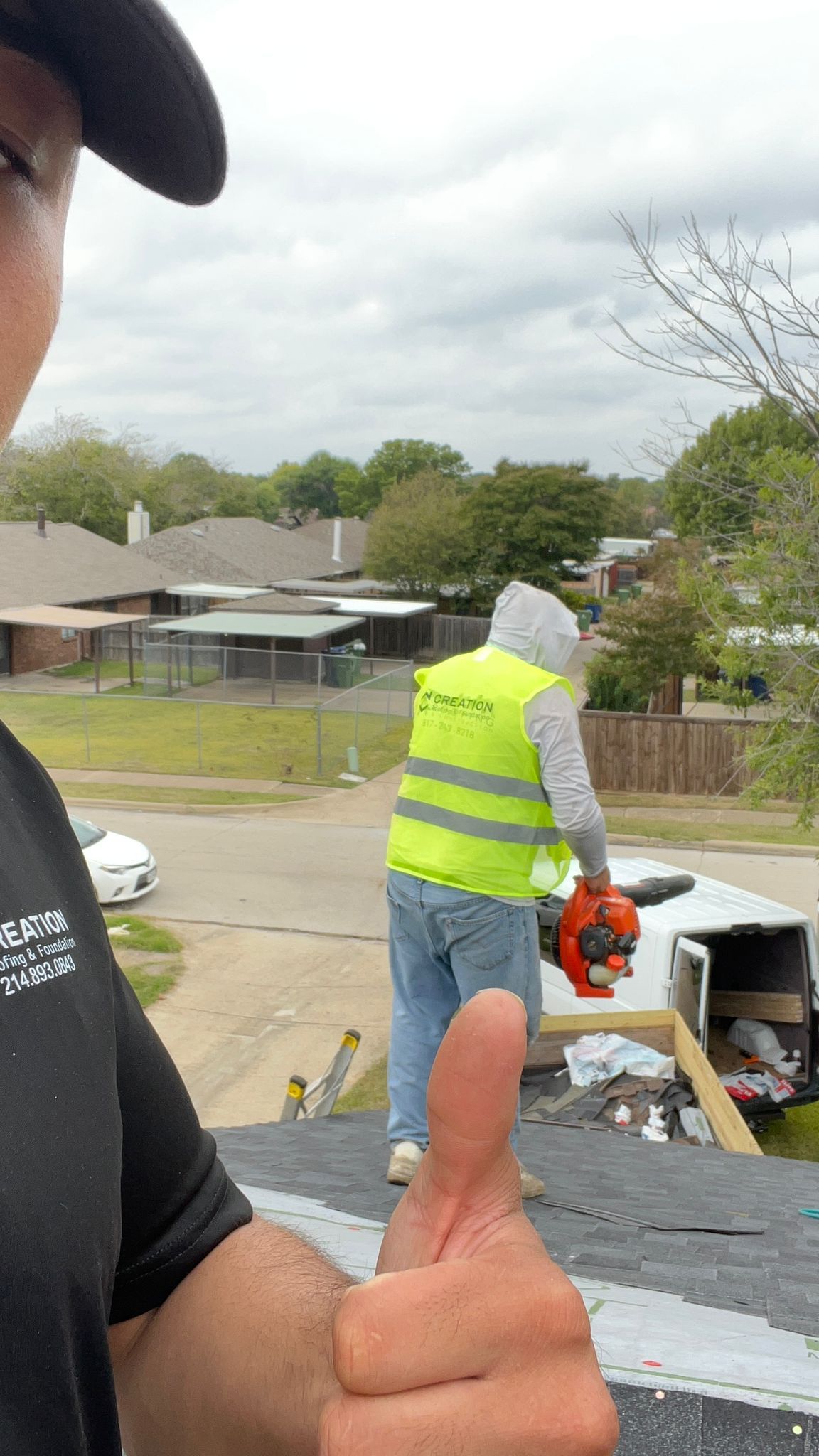 Person giving thumbs up on roof as worker in vest uses blower. Suburban setting.