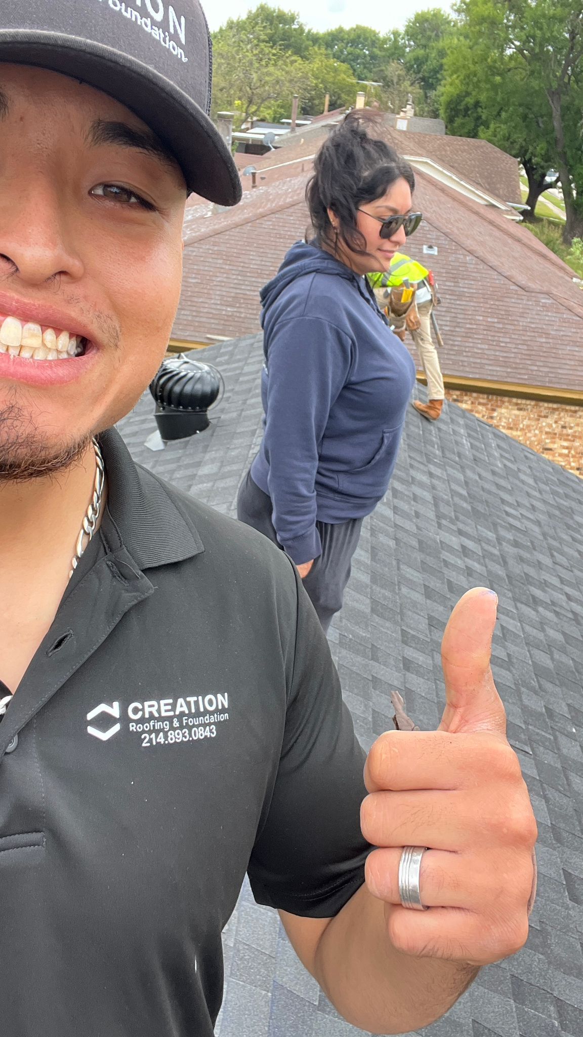 Man on roof giving thumbs up, woman and worker in background. Roof appears to be under construction, overcast day.