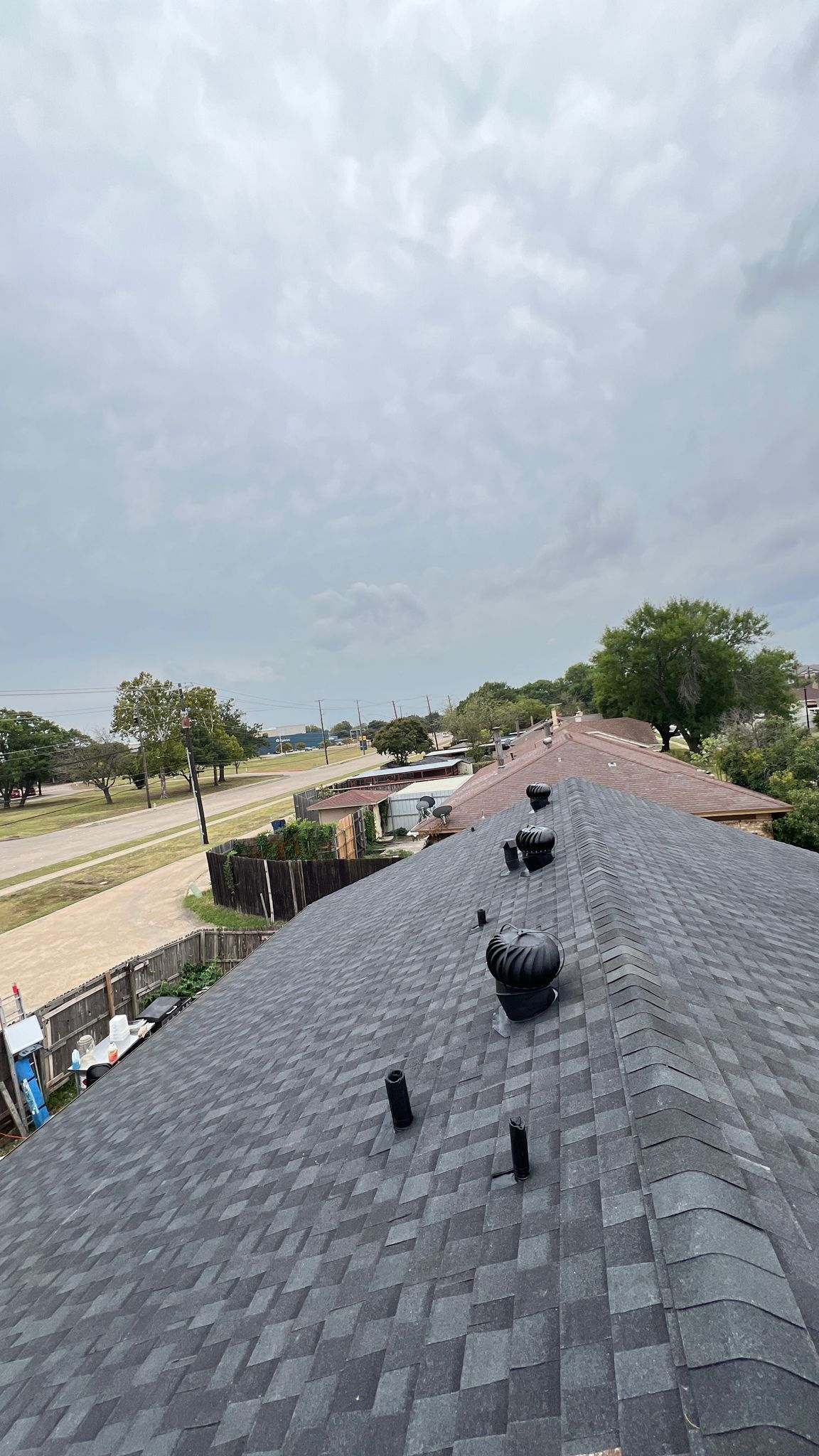 View of a dark asphalt shingle roof, with vents, looking towards a road and cloudy sky.