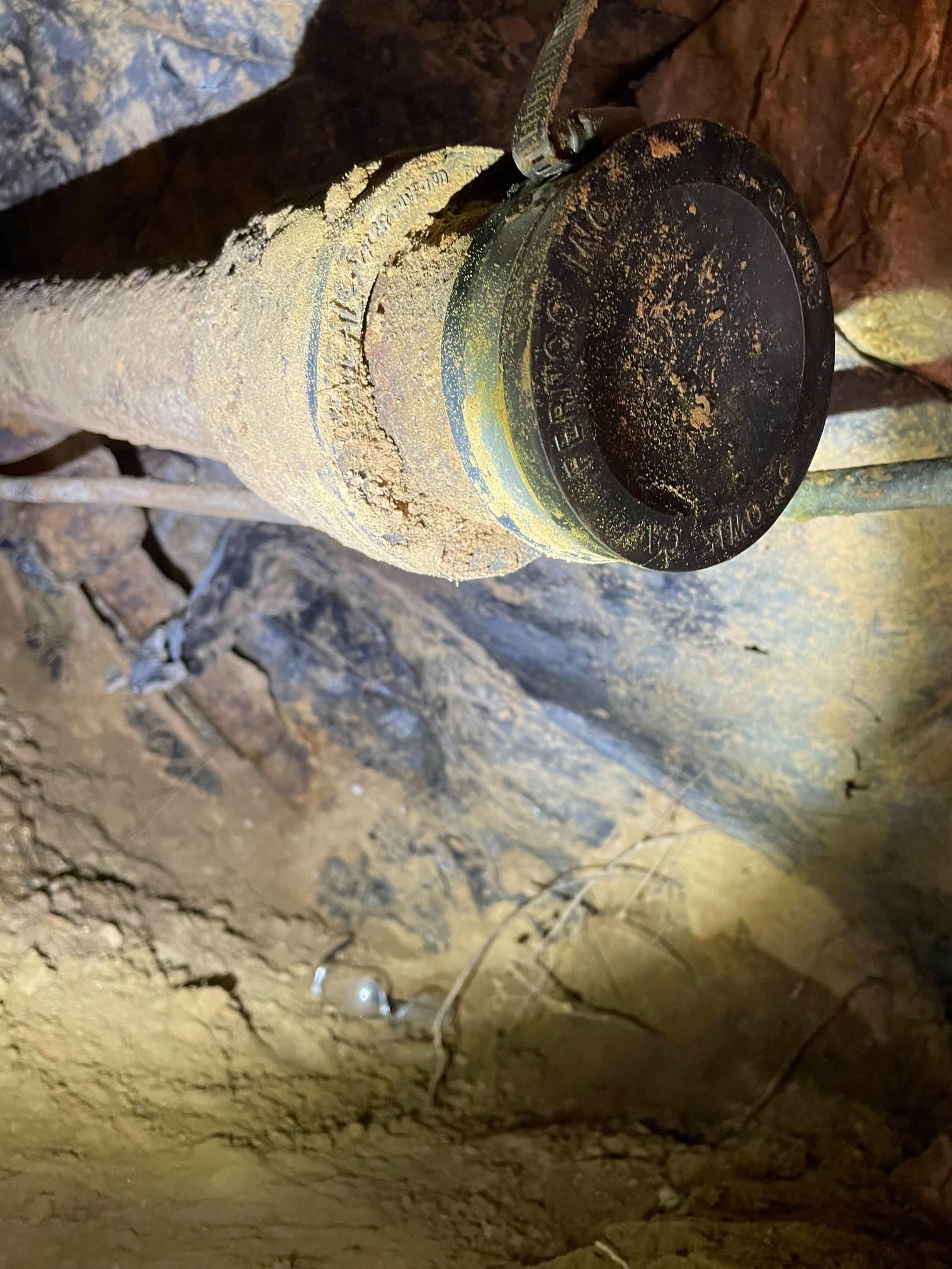 Rusty pipe in a dark, earthy environment, possibly a crawl space or underground.