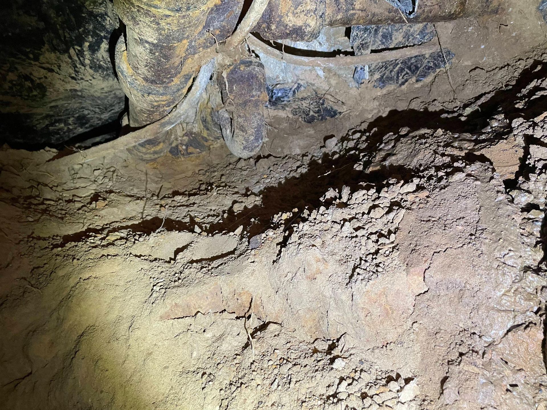 Interior view of a dark tunnel. Rusty pipes are visible overhead, and the walls are covered in dirt and debris.