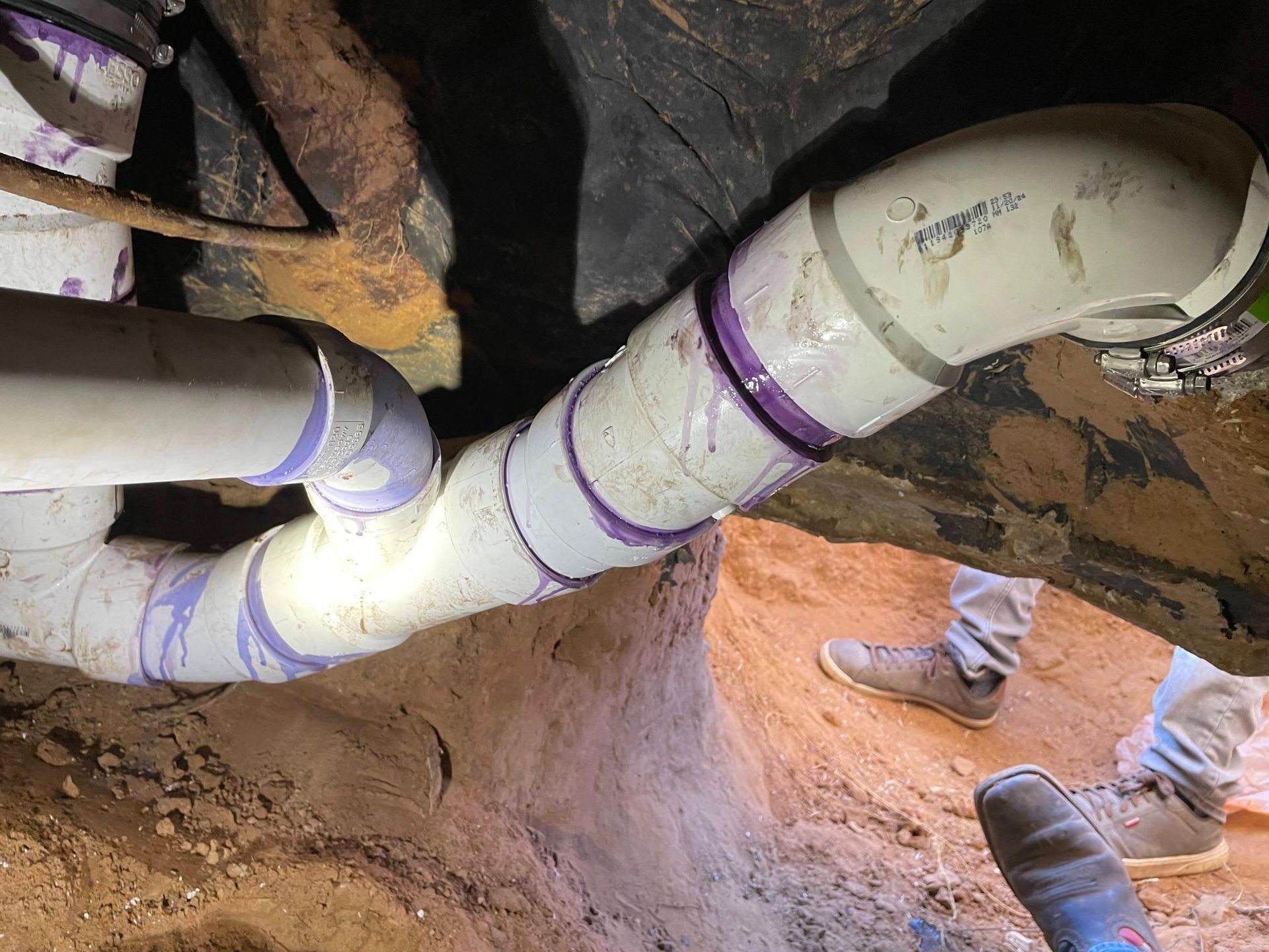 Plumbing pipes with purple primer are in a dirt trench, near a person's legs.