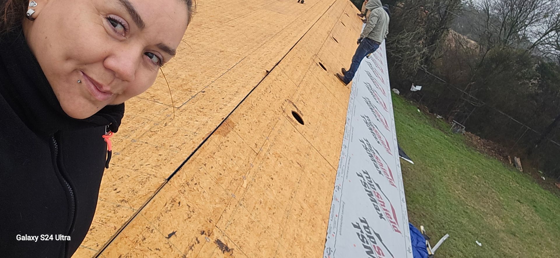 Woman smiling on a slanted roof with other person working. The roof is partially covered in wood and tarpaper.
