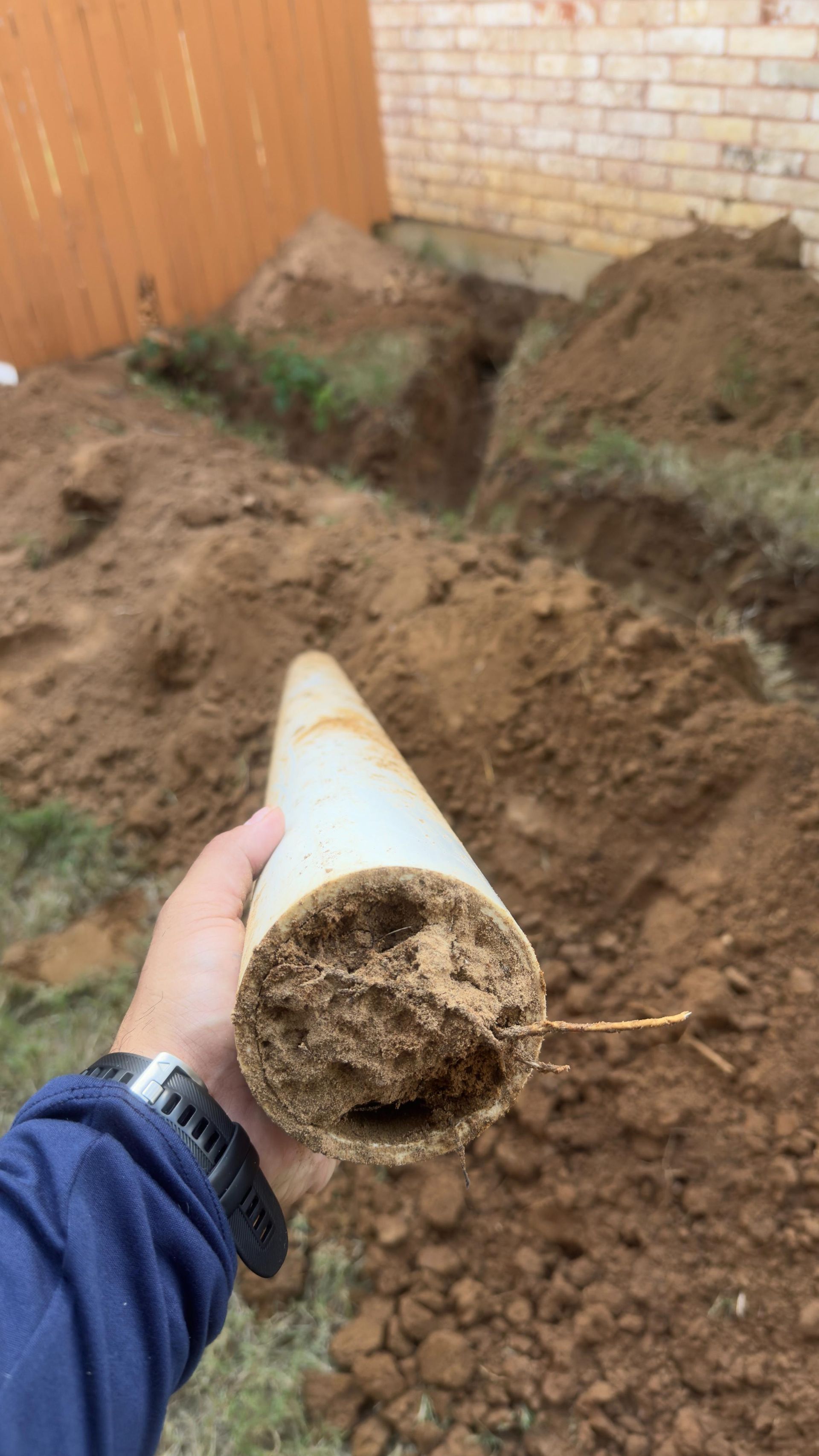 A hand holding a section of corroded white pipe. Dirt fills the pipe’s interior, with a trench in the background.