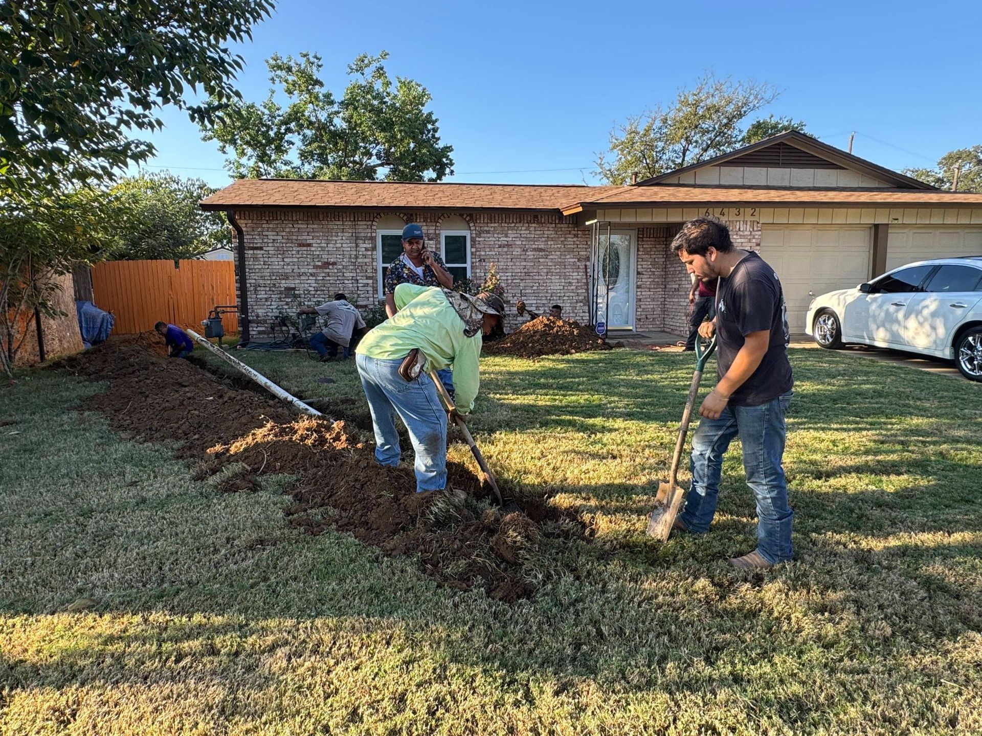 People digging trench in a yard, near a house with brown siding and a white door. A white car is parked nearby.