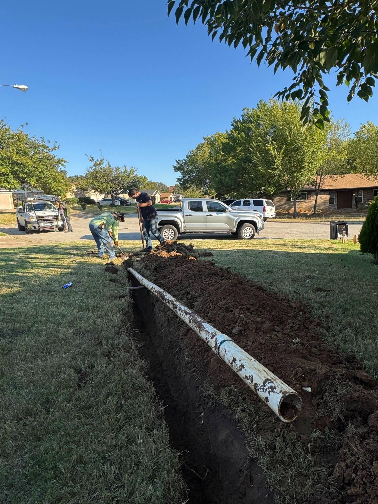 Workers installing a white PVC pipe in a trench on a grassy lawn with a truck parked nearby. Sunny day.