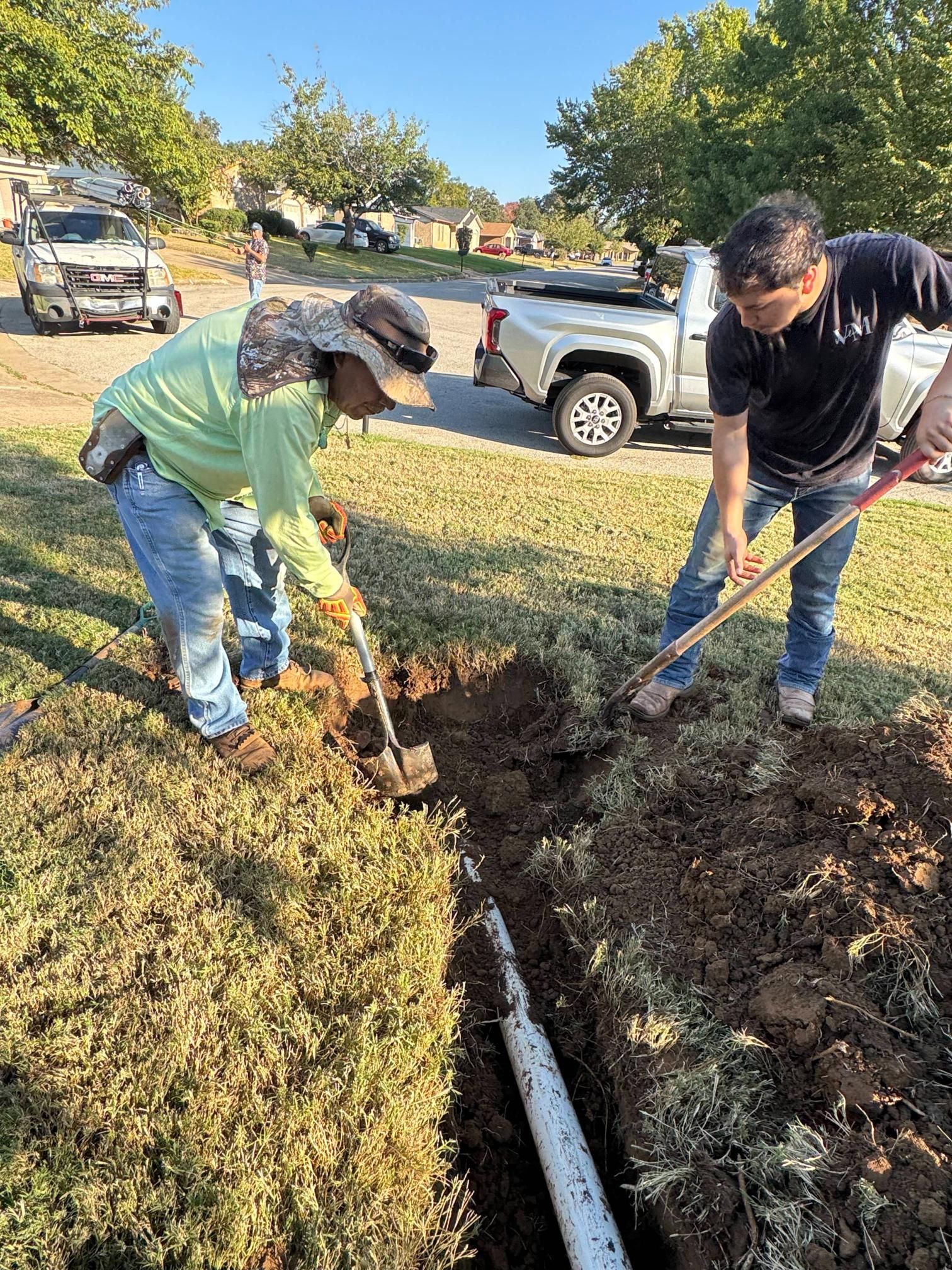Two people digging a trench in a grassy area next to a white pipe. A truck and trees are visible.