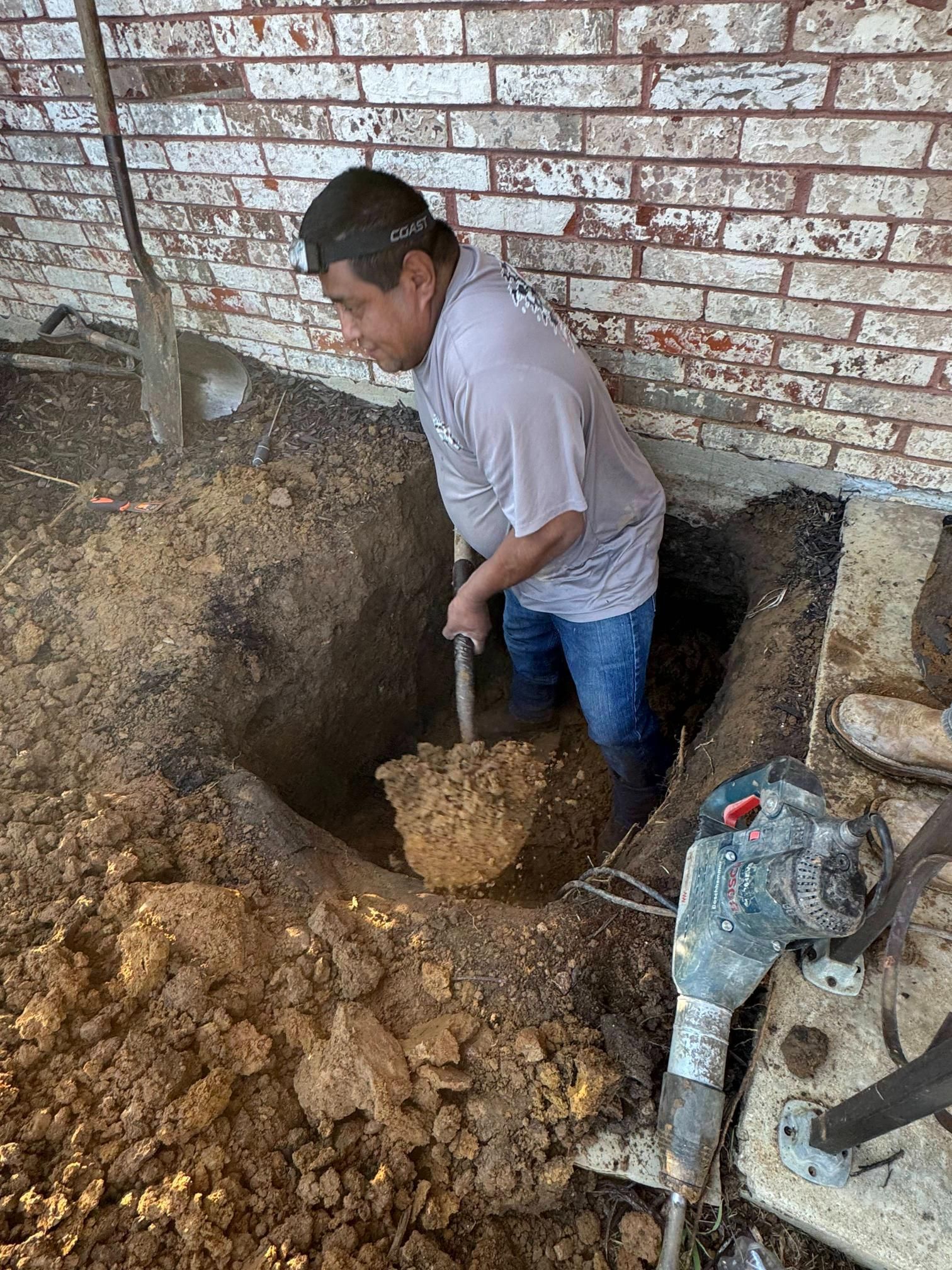 Man digging in a hole near a brick wall with a shovel.
