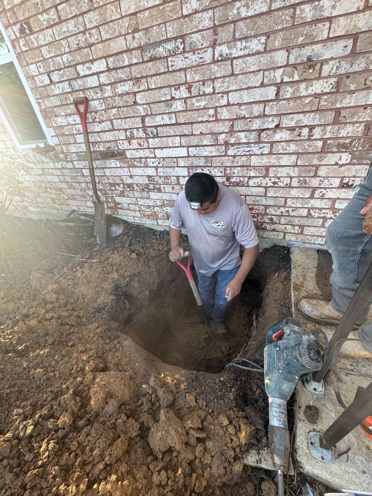 Person digging in dirt next to a brick building. A shovel and jackhammer are present.