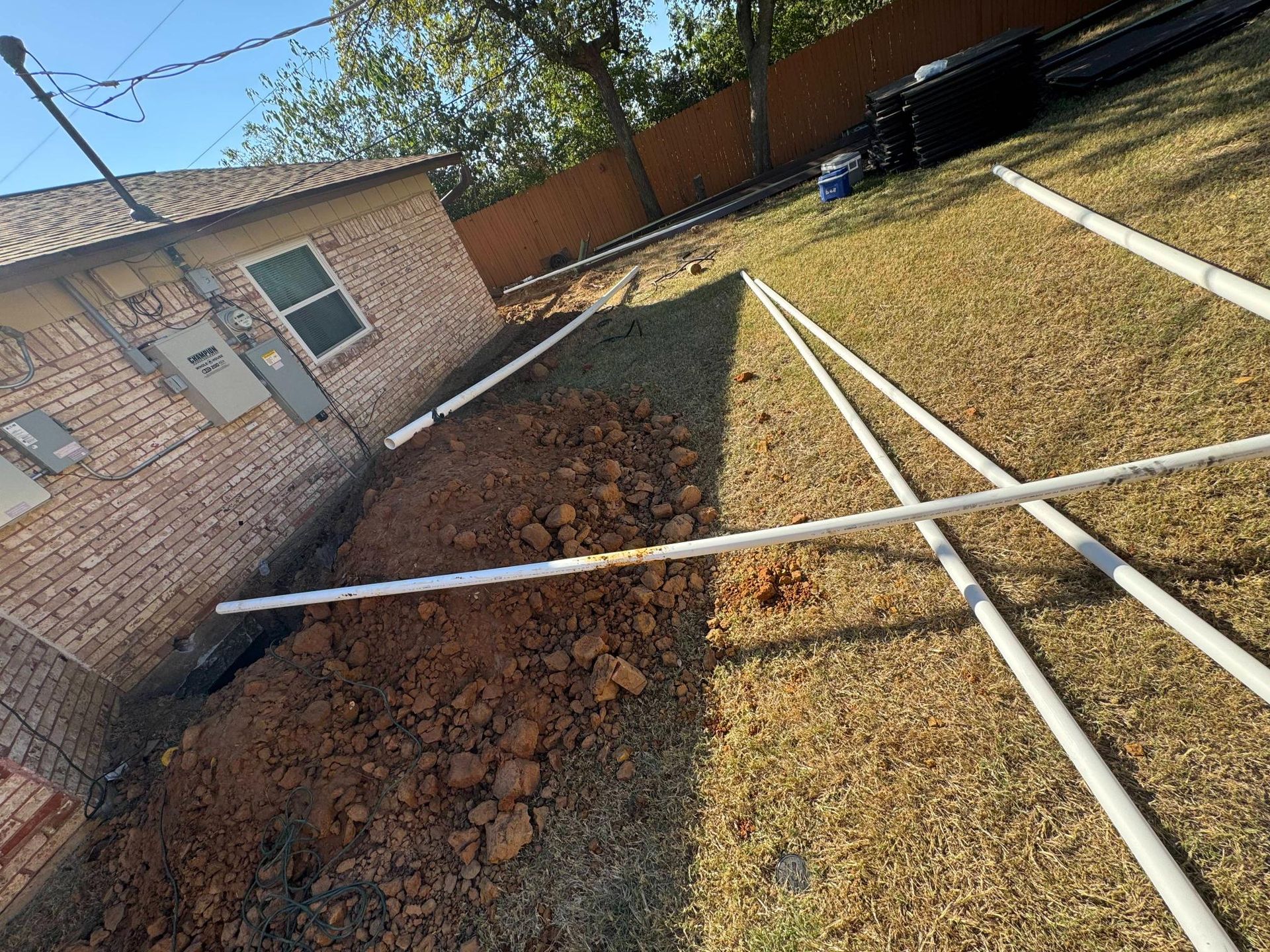 Backyard with dug trench and white PVC pipes laid out near a brick building and brown fence.