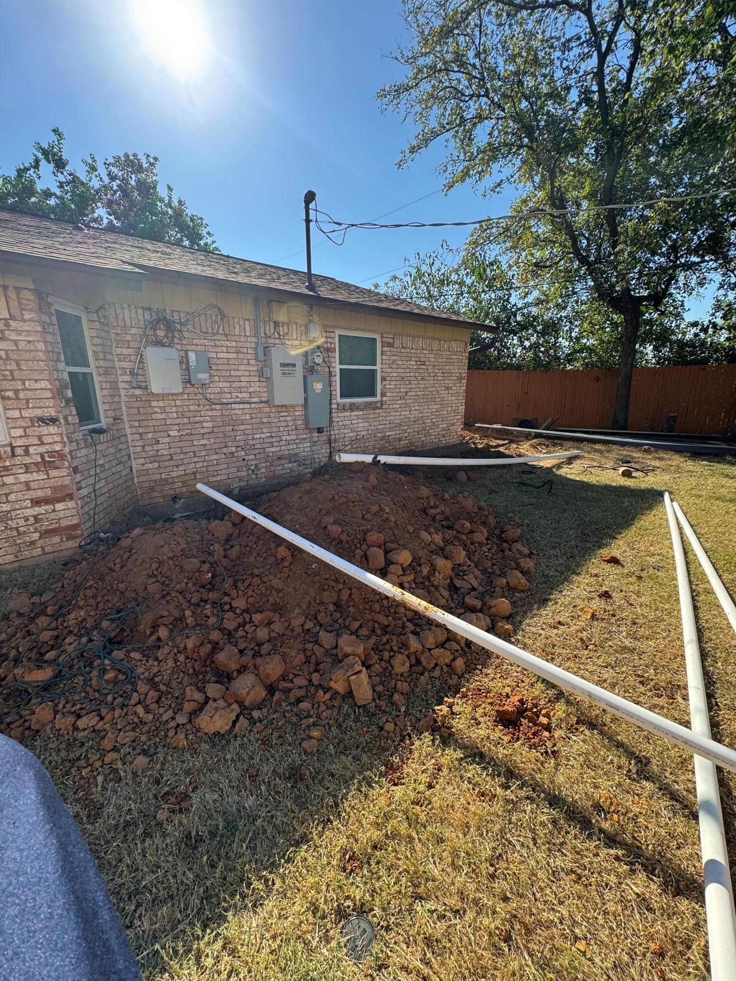 Backyard construction with exposed white pipes and a brick building under a sunny sky.