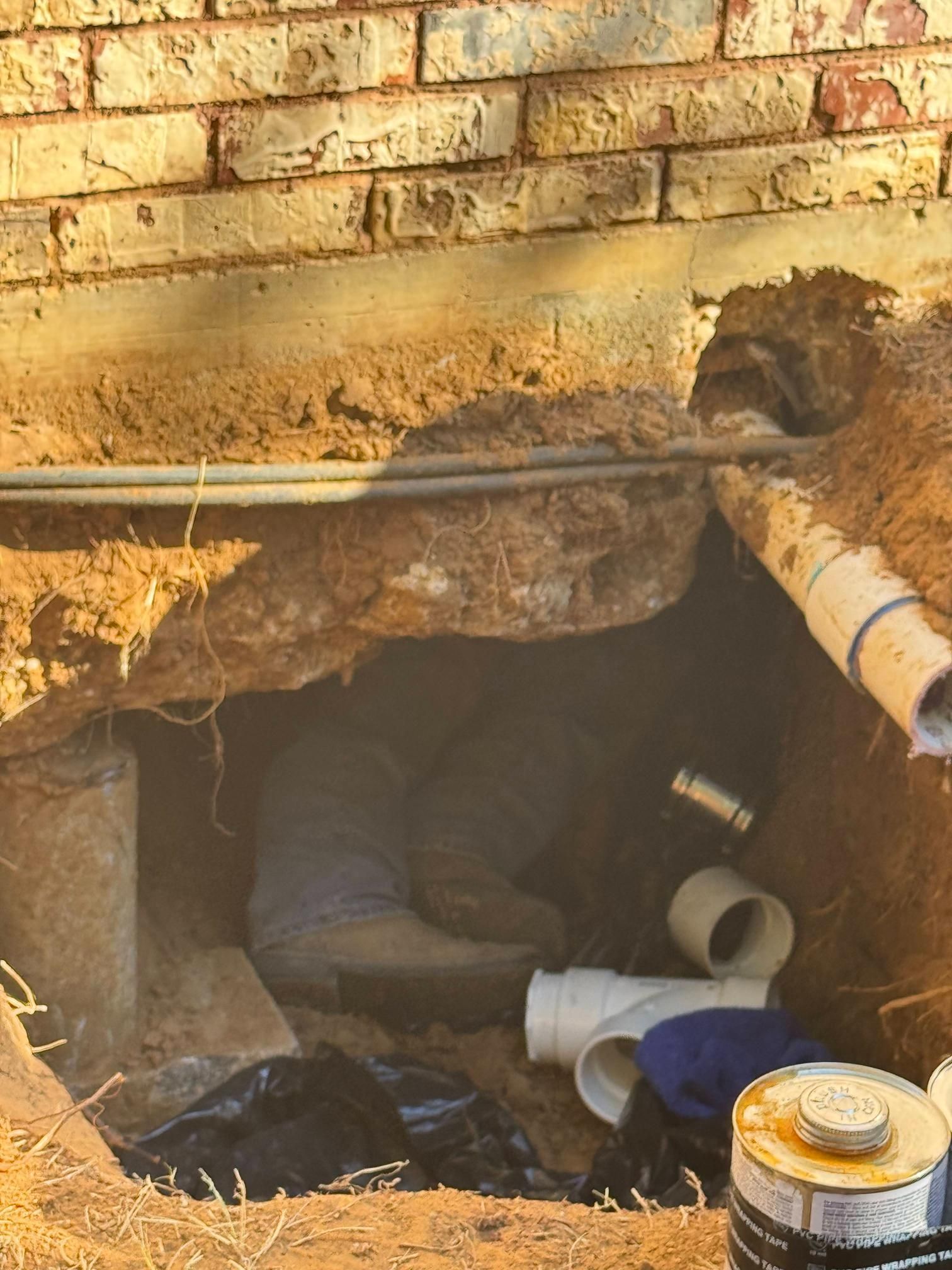 Person working on plumbing pipes under a brick building, visible in an excavated hole.