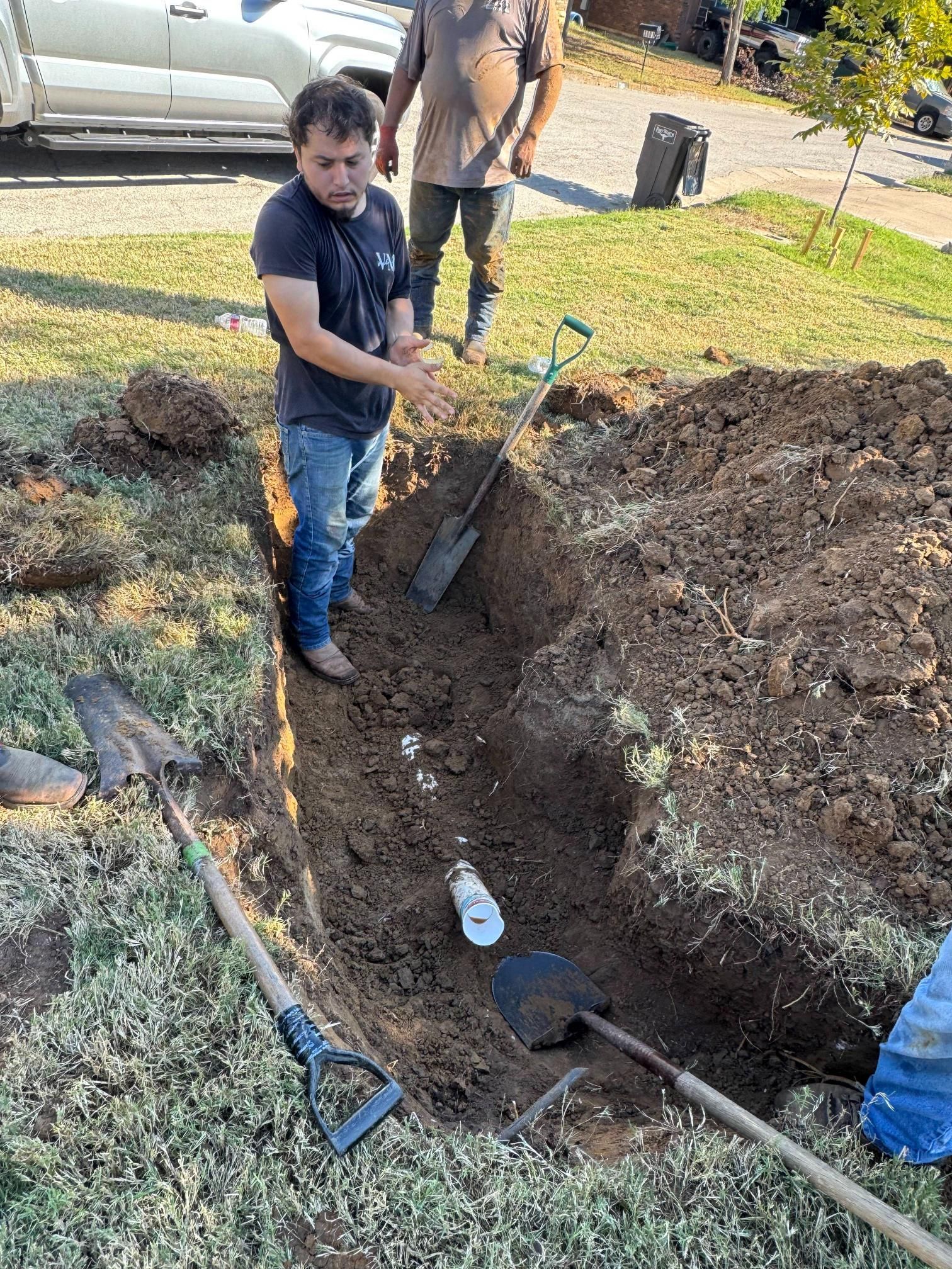 Two men in a trench, one points at a pipe. Shovels are present, outdoors.