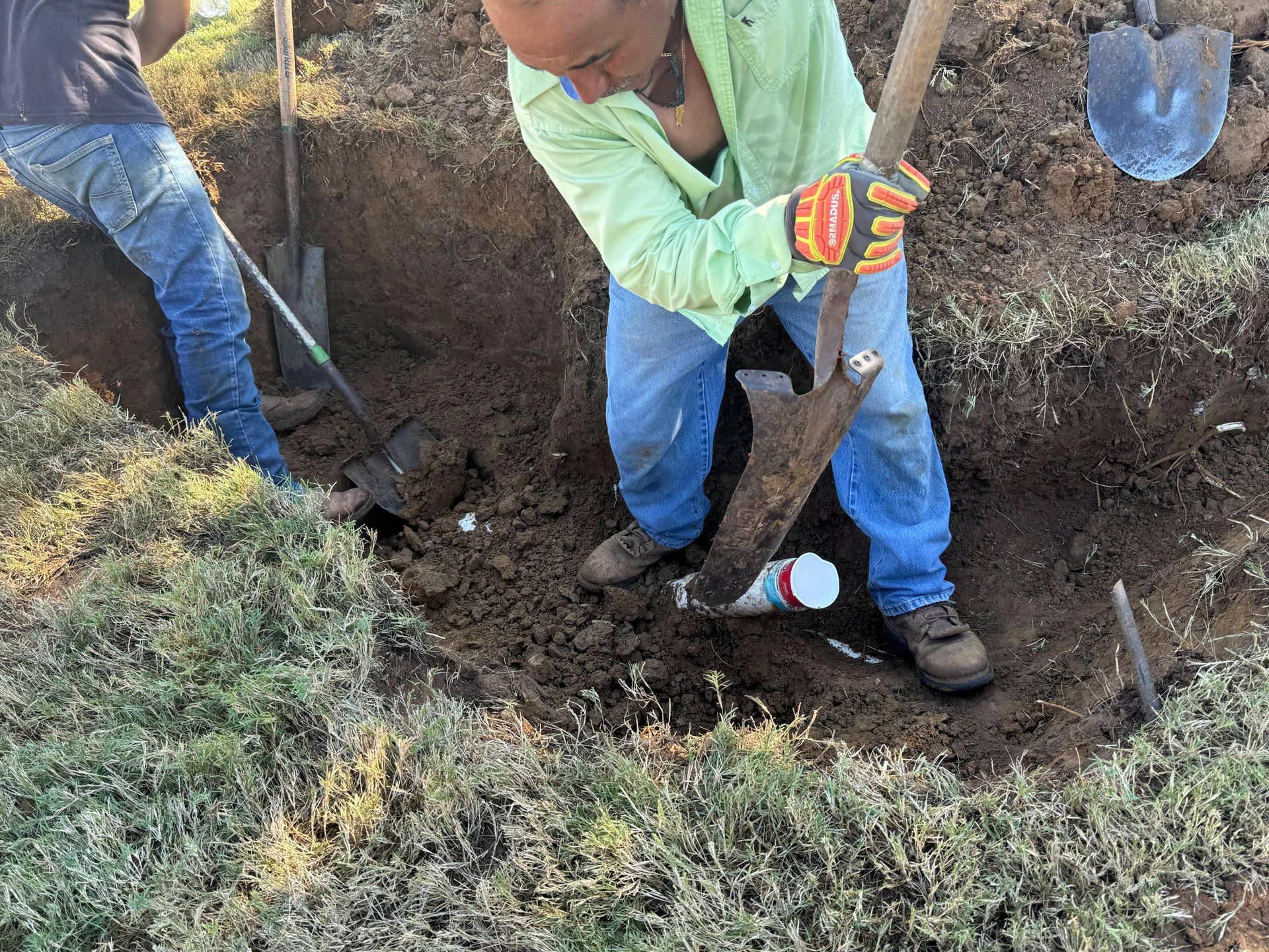 Two people digging in a trench near a white pipe. One uses a shovel, the other a spade.