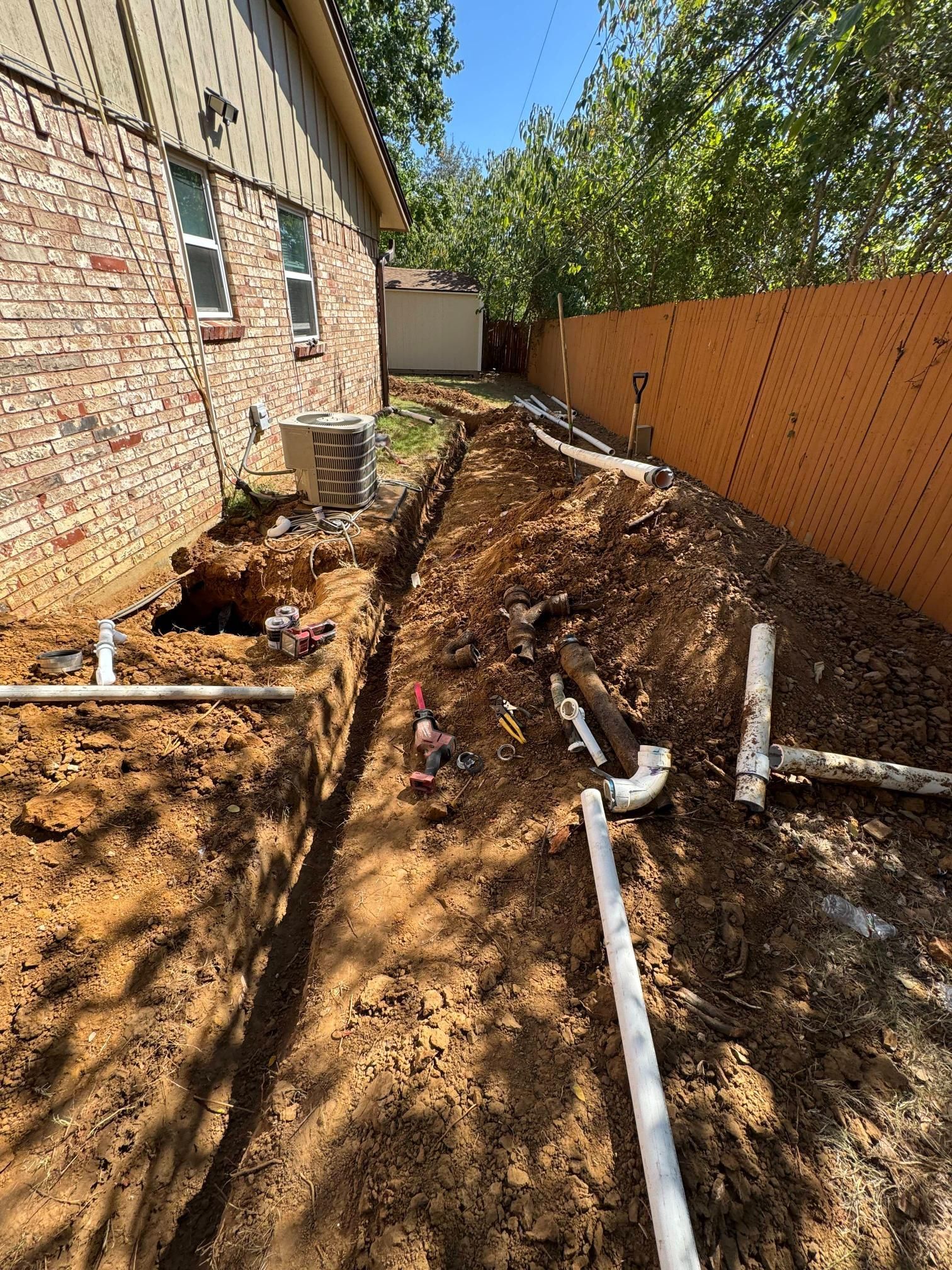 A trench dug next to a brick building, with exposed pipes and soil. Orange fence in background.