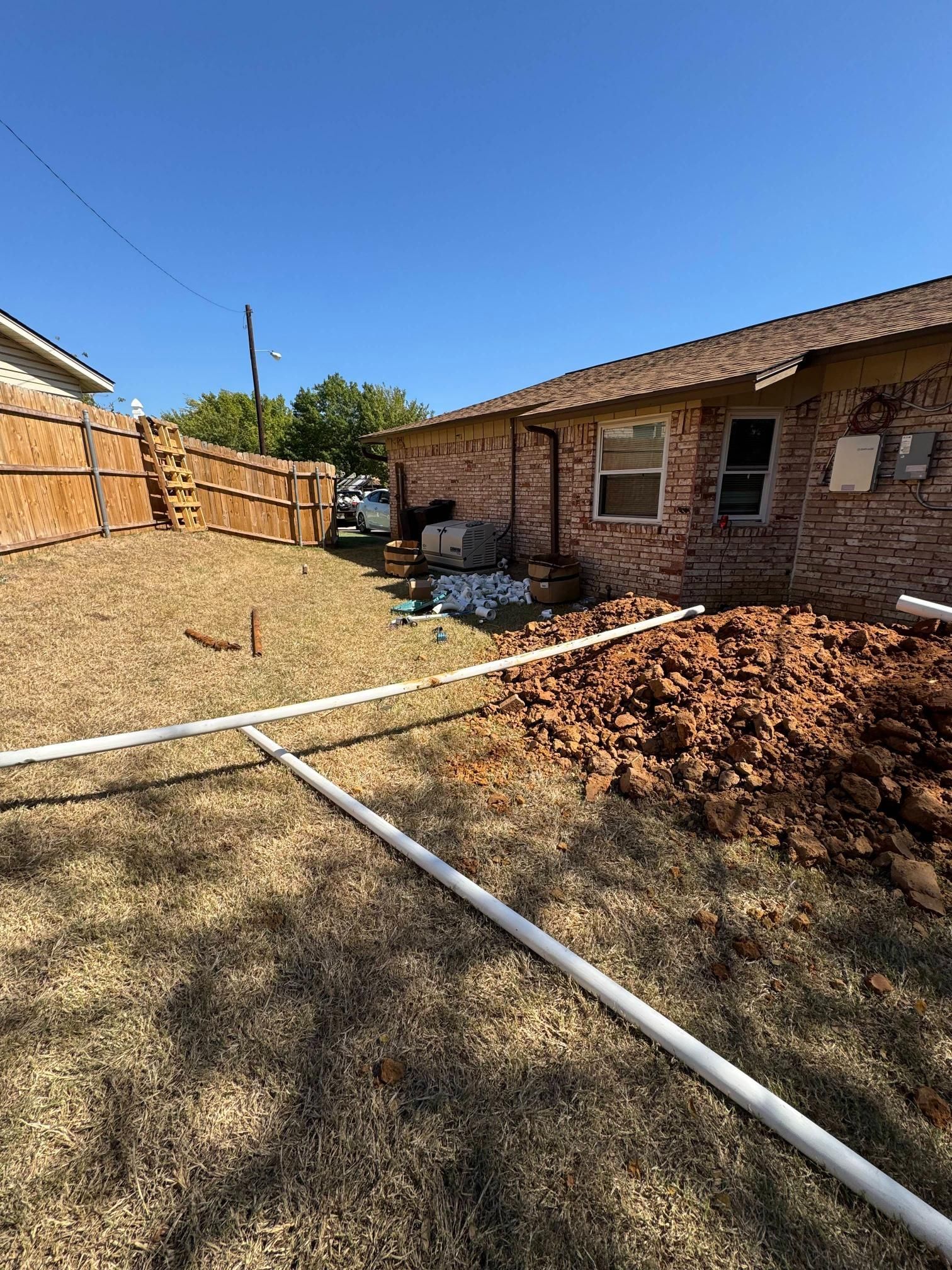 Backyard with exposed pipes, dirt pile, and brick house under a blue sky.