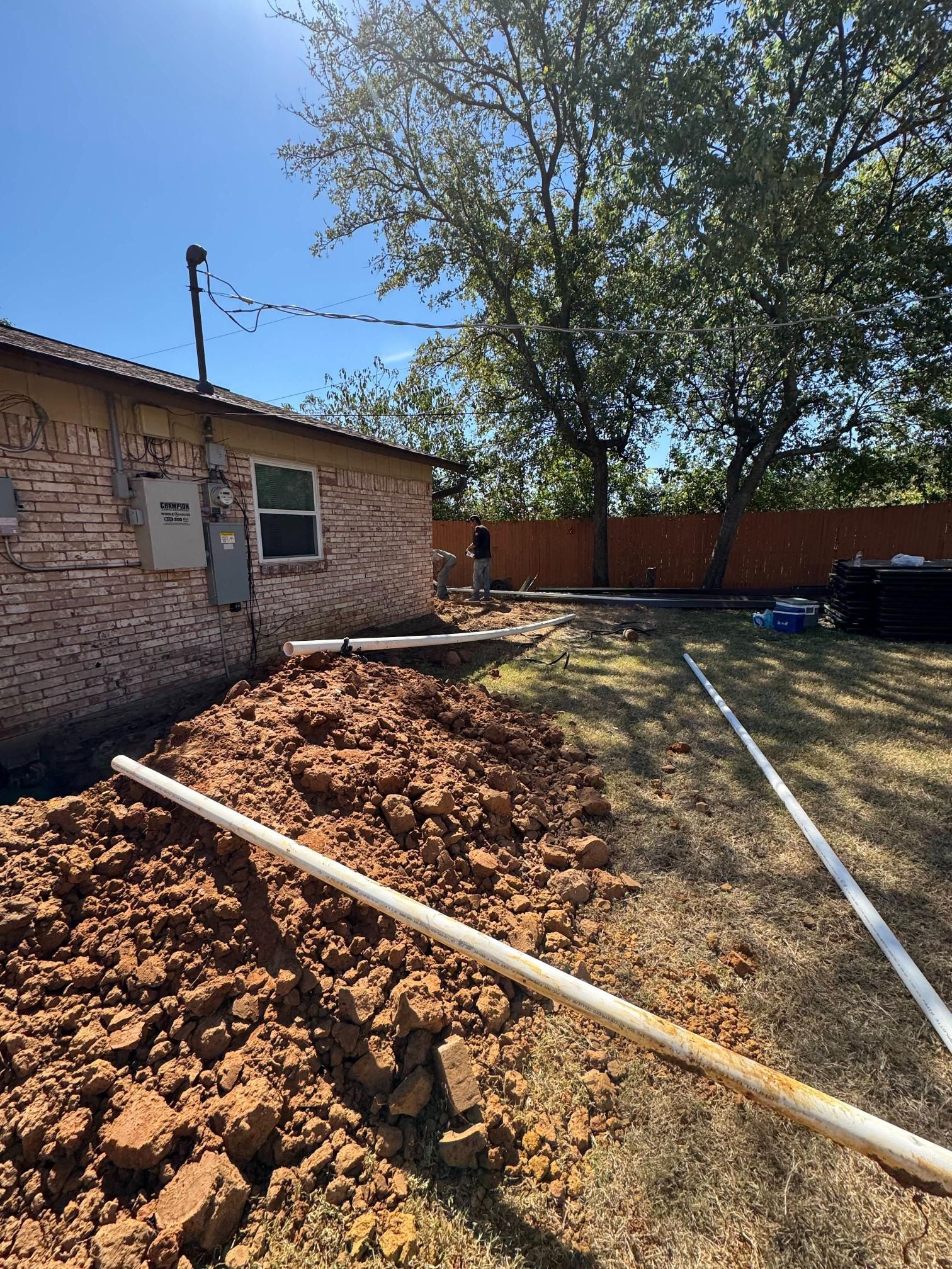 Backyard with a trench, pipe, and pile of dirt near a brick building and fence on a sunny day.