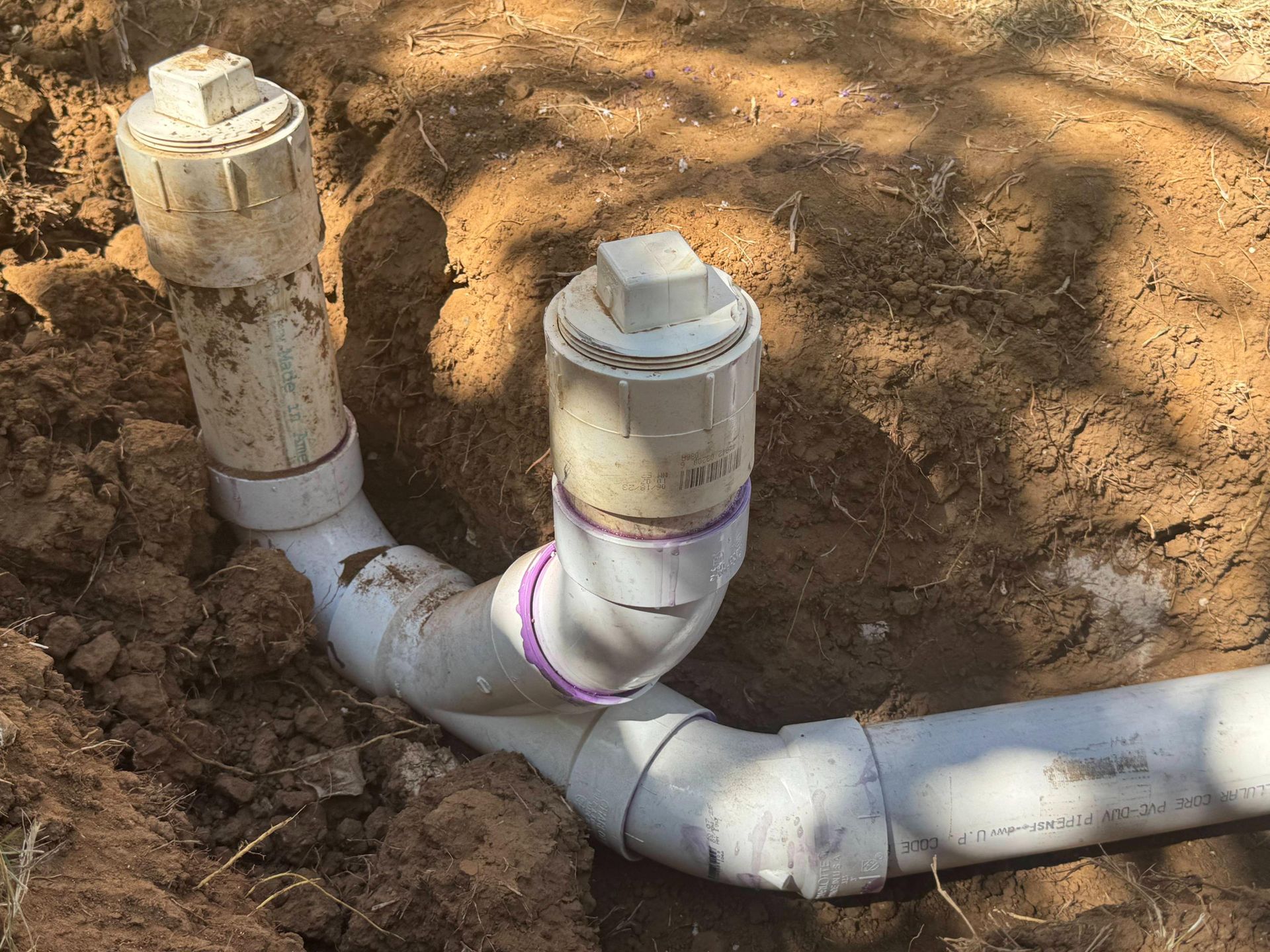 White PVC pipes in a dirt trench, connected with elbows. Two capped vertical pipes are visible.
