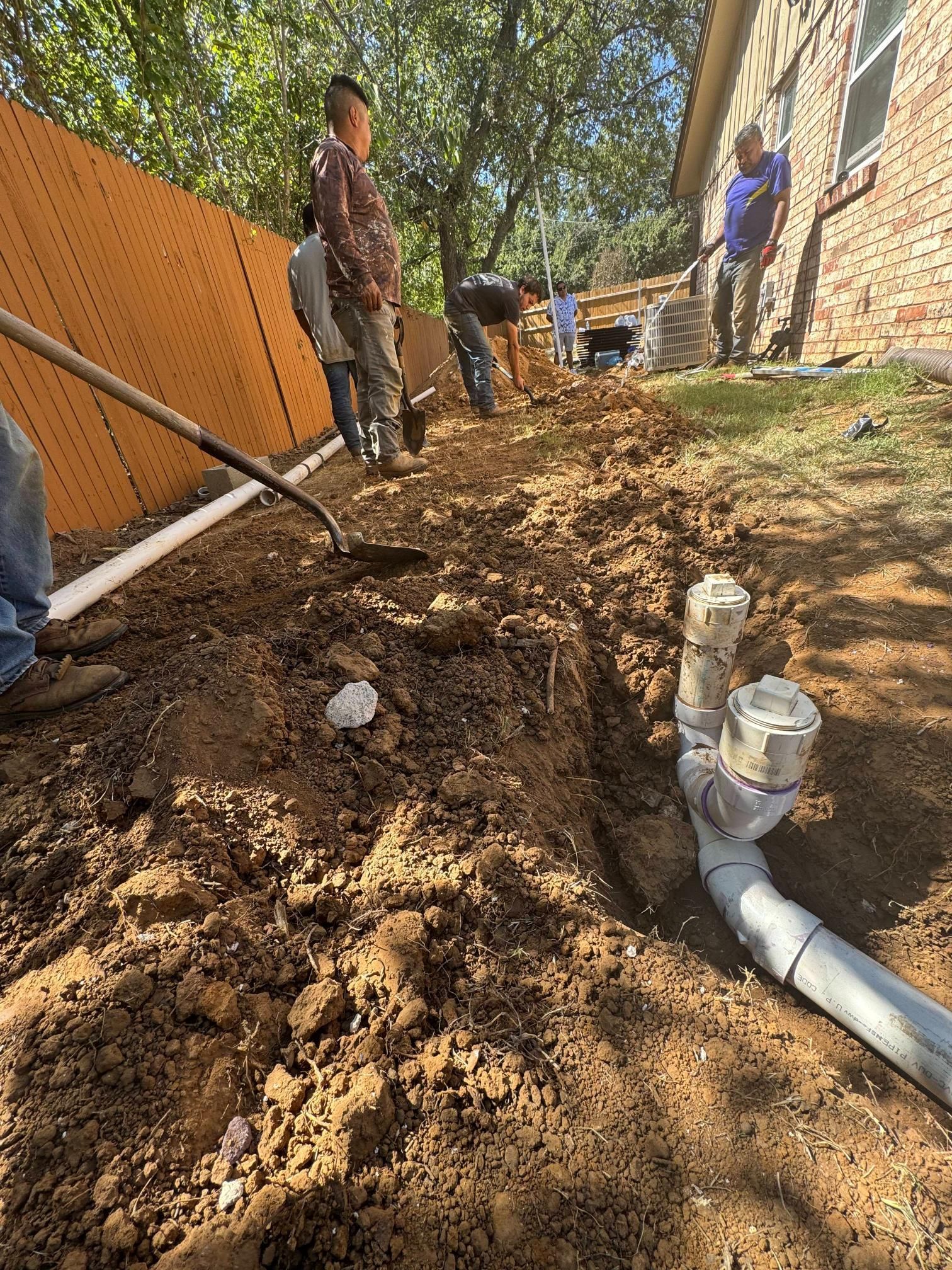 Workers digging trenches and installing pipes near a house and fence.