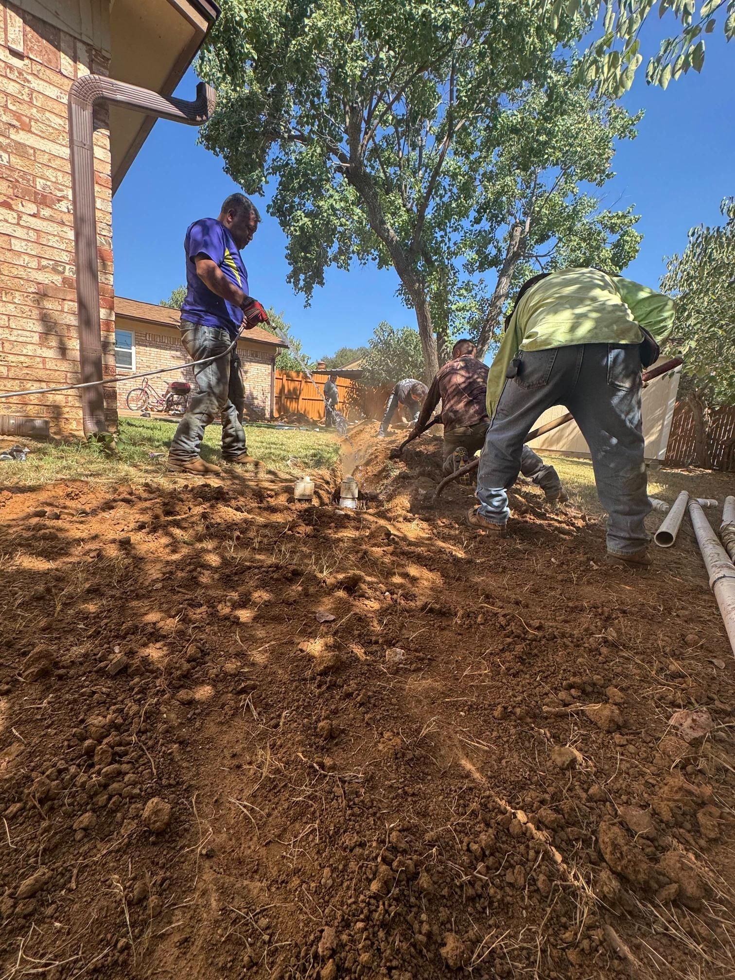 Workers digging in a yard near a brick house. One stands, two kneel, working. Brown dirt and green trees visible.