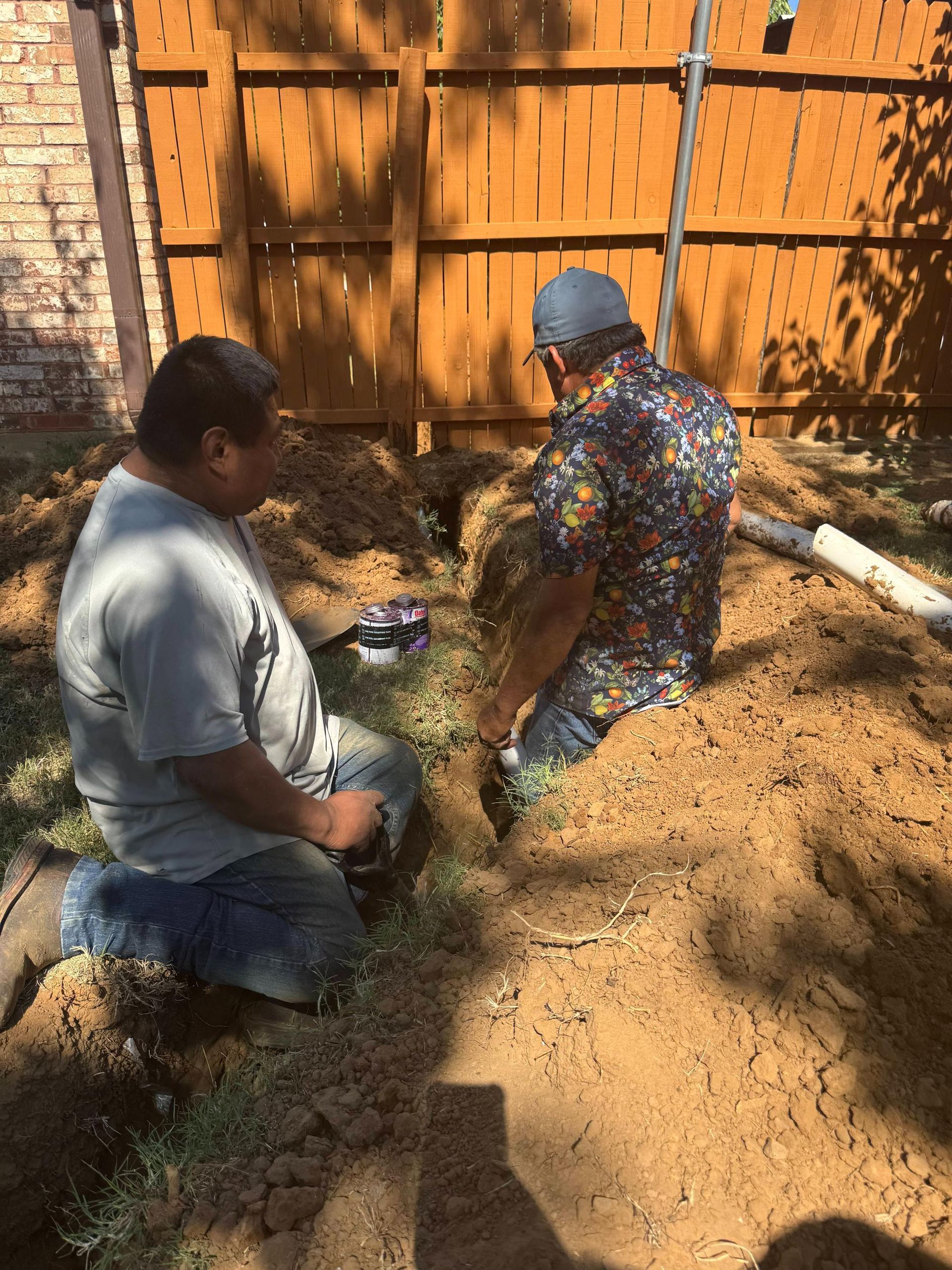 Two people digging a trench in a yard near a wooden fence.