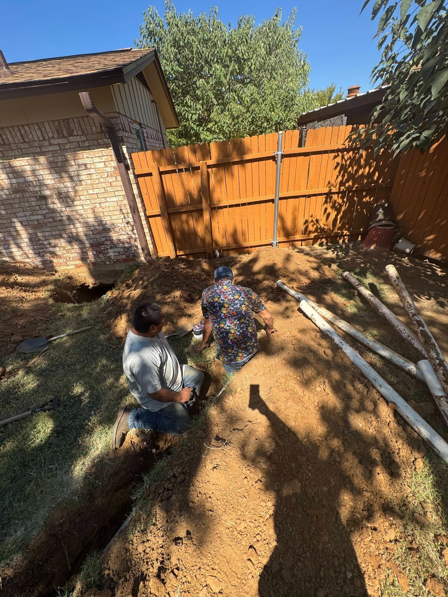 Two people digging trenches in a yard near a wooden fence and a brick building, under a sunny sky.