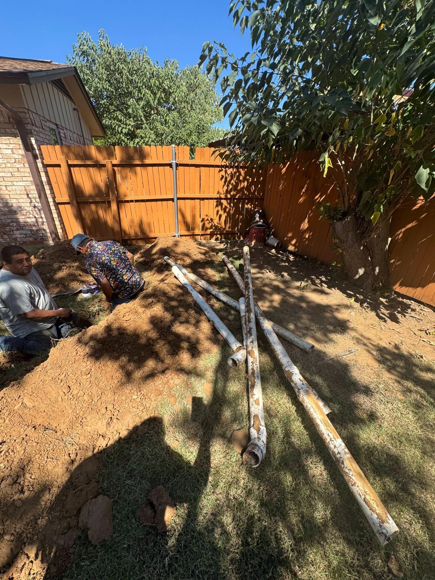 Two people digging in a yard near a fence, with several pipes laying on the ground.