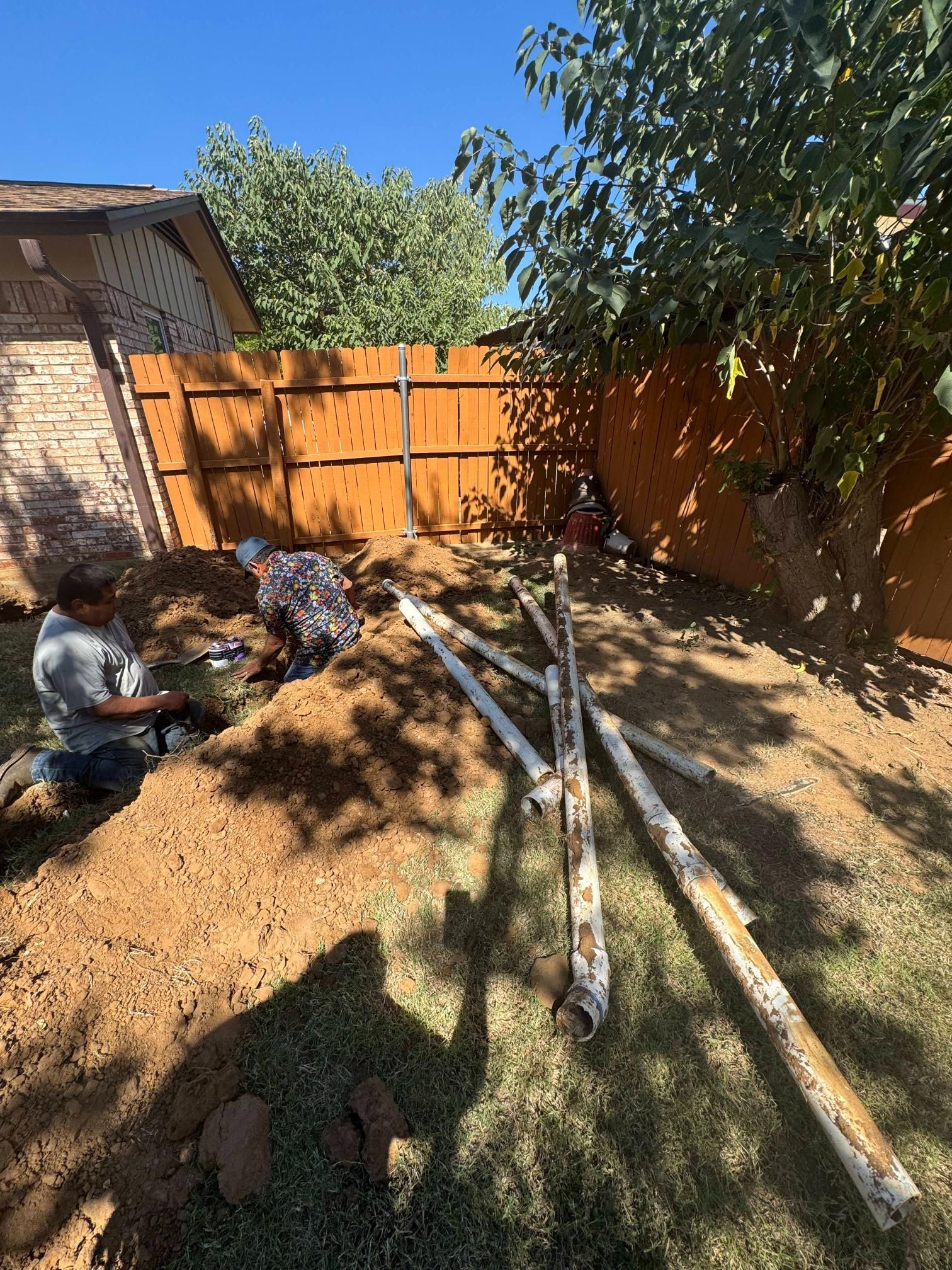 Workers installing pipes in a yard. Trenches are dug in the dirt. White PVC pipes lie nearby fence.
