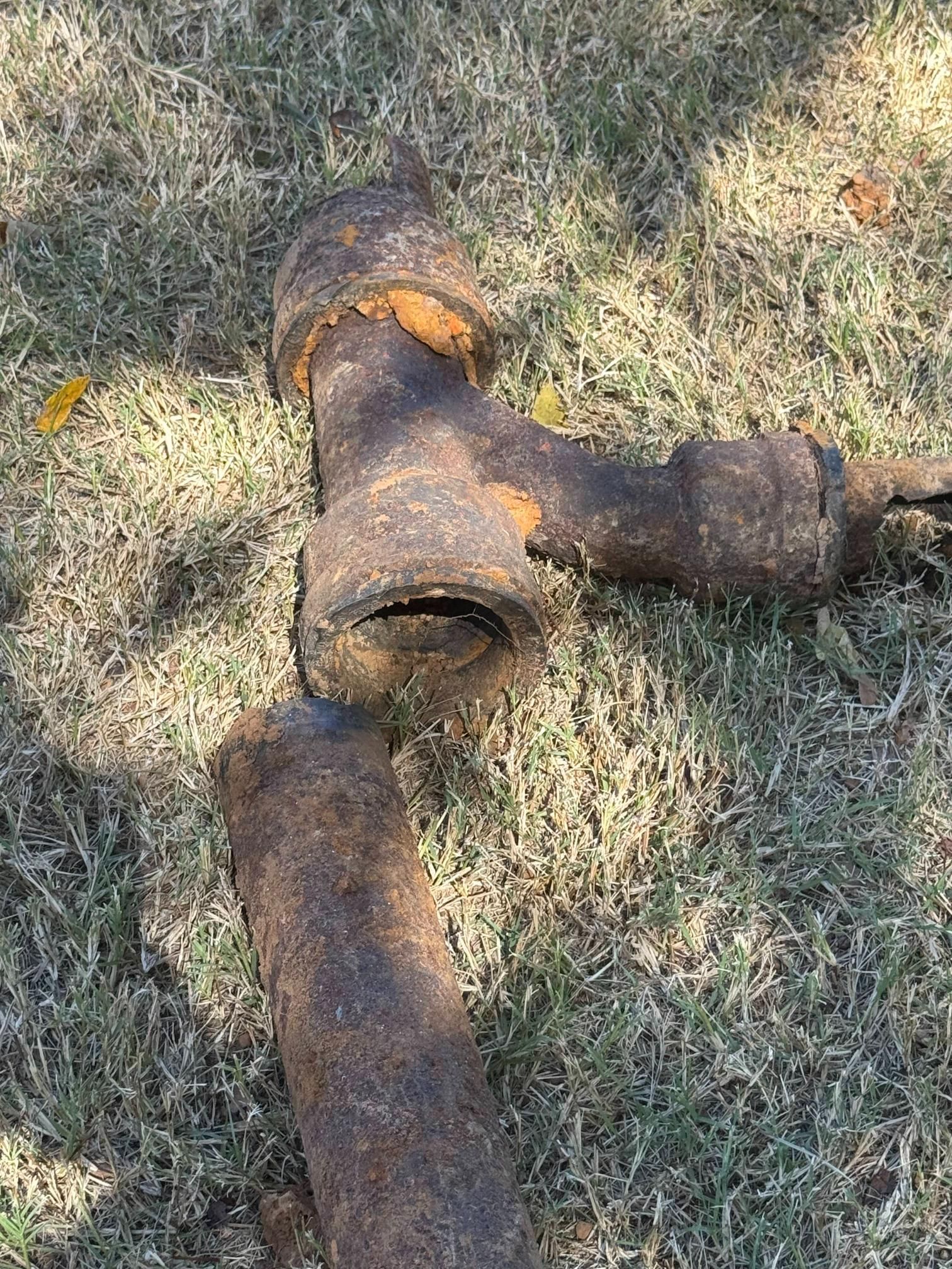 Rusty, broken iron pipe fitting on grass. A large T-shaped piece with a section of pipe detached. Brown and orange.