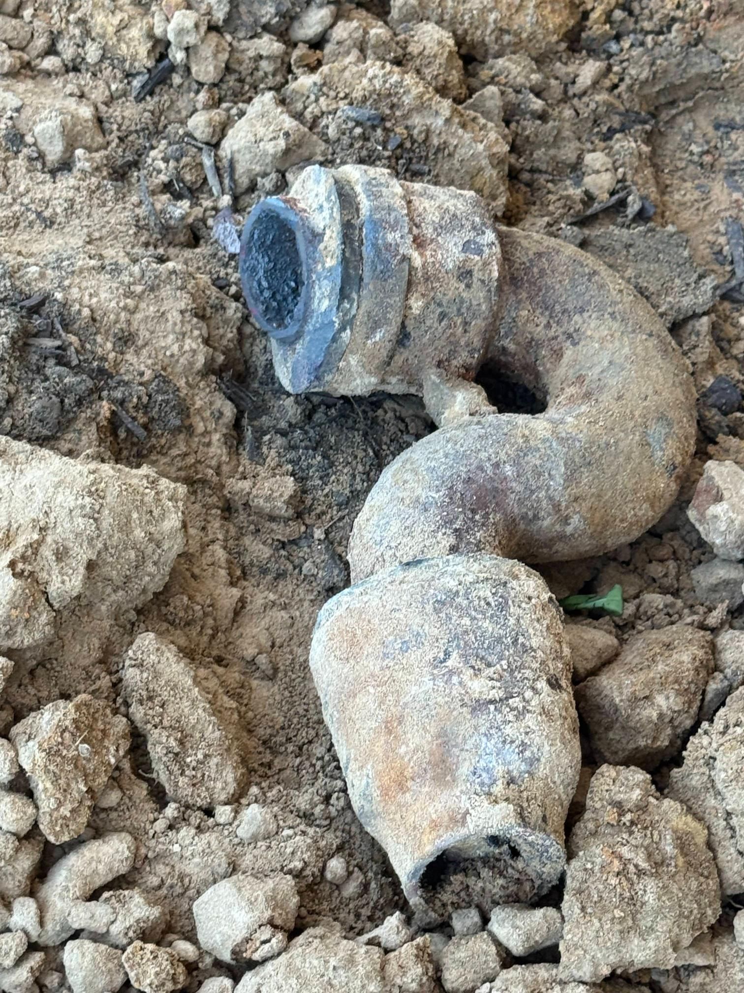 A curved, buried pipe fitting, possibly metal, in loose dirt and rocks.