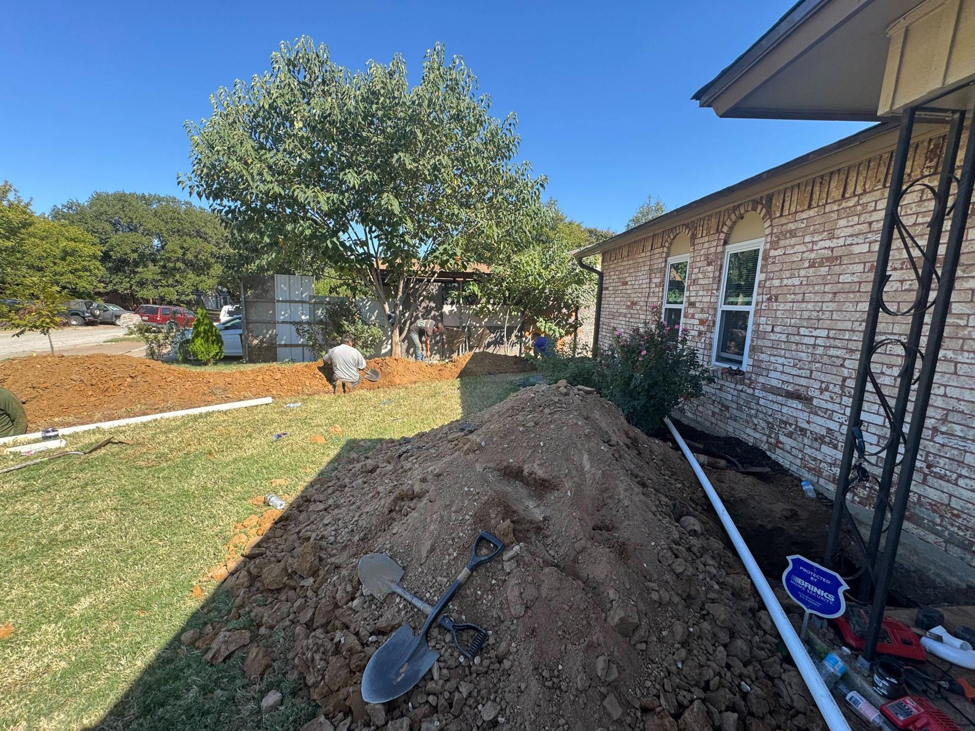 Trenches dug in a yard next to a brick house. Soil piles, shovel, and piping are visible.