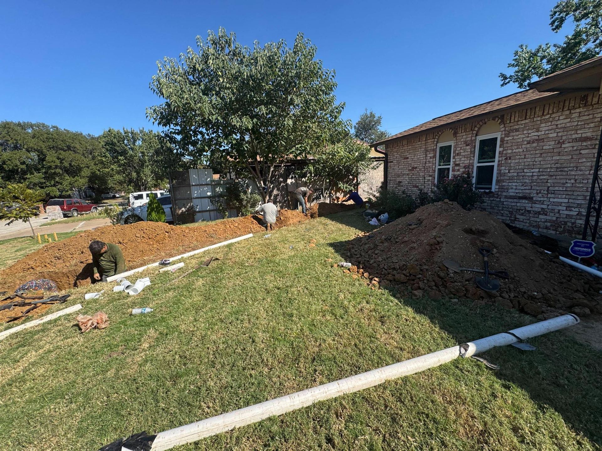 Yard with dug trenches, piles of dirt, and white pipes, likely for plumbing work next to a brick house.