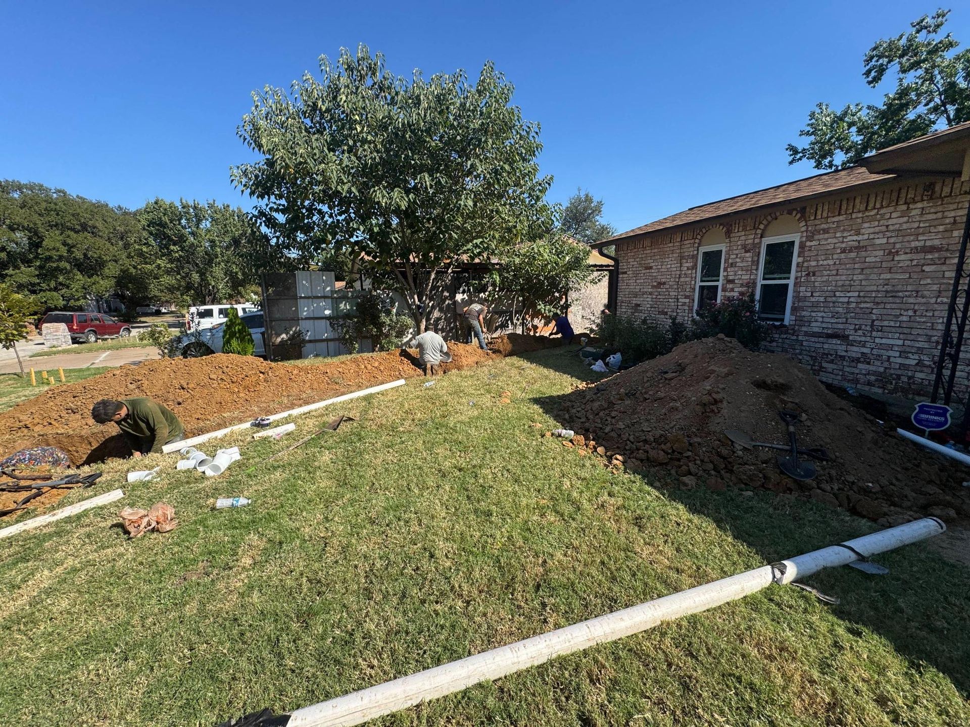 Workers installing white pipes in a grassy yard, near a brick house and tree. Dirt piles are present.