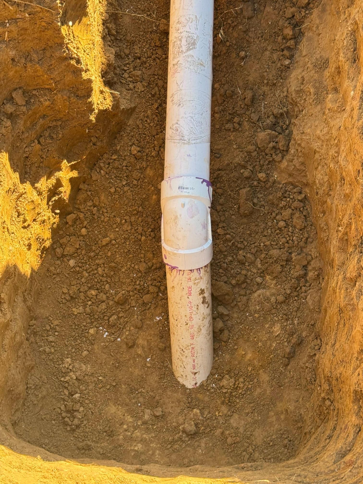 White PVC pipe section with a coupling joint, set in a brown soil trench.