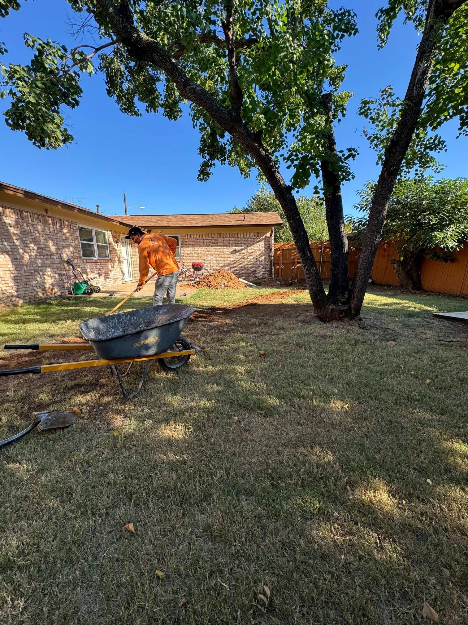 Person shoveling dirt in a yard near a tree, wheelbarrow in the foreground, brick building in the background.
