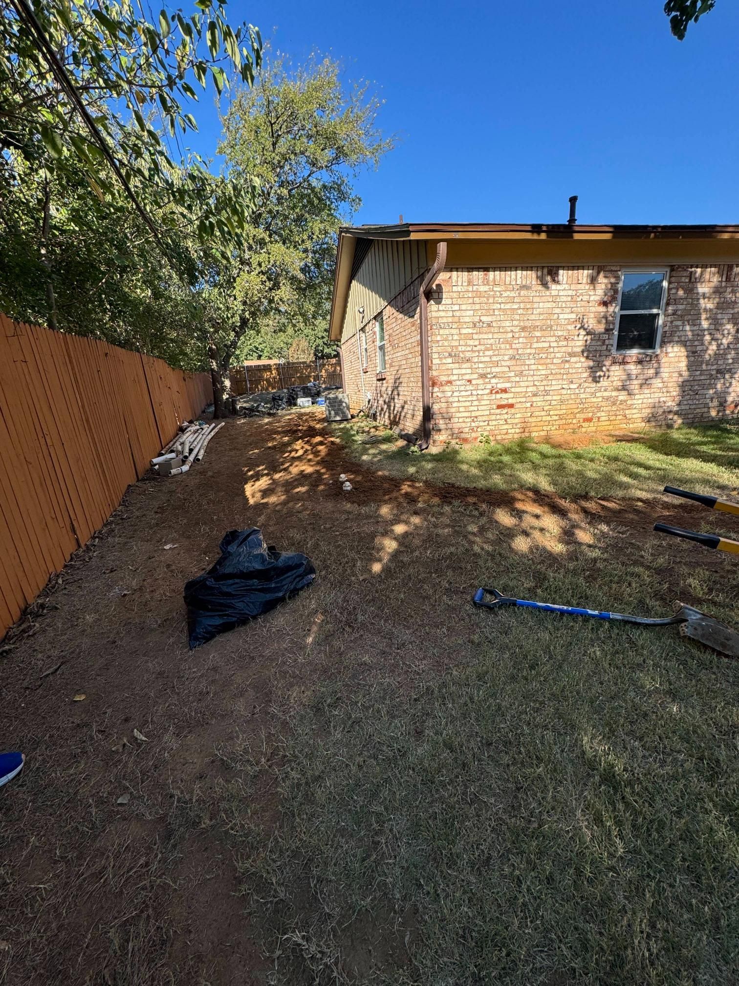 A backyard with a brown fence, dirt patch, and a brick house under a blue sky.