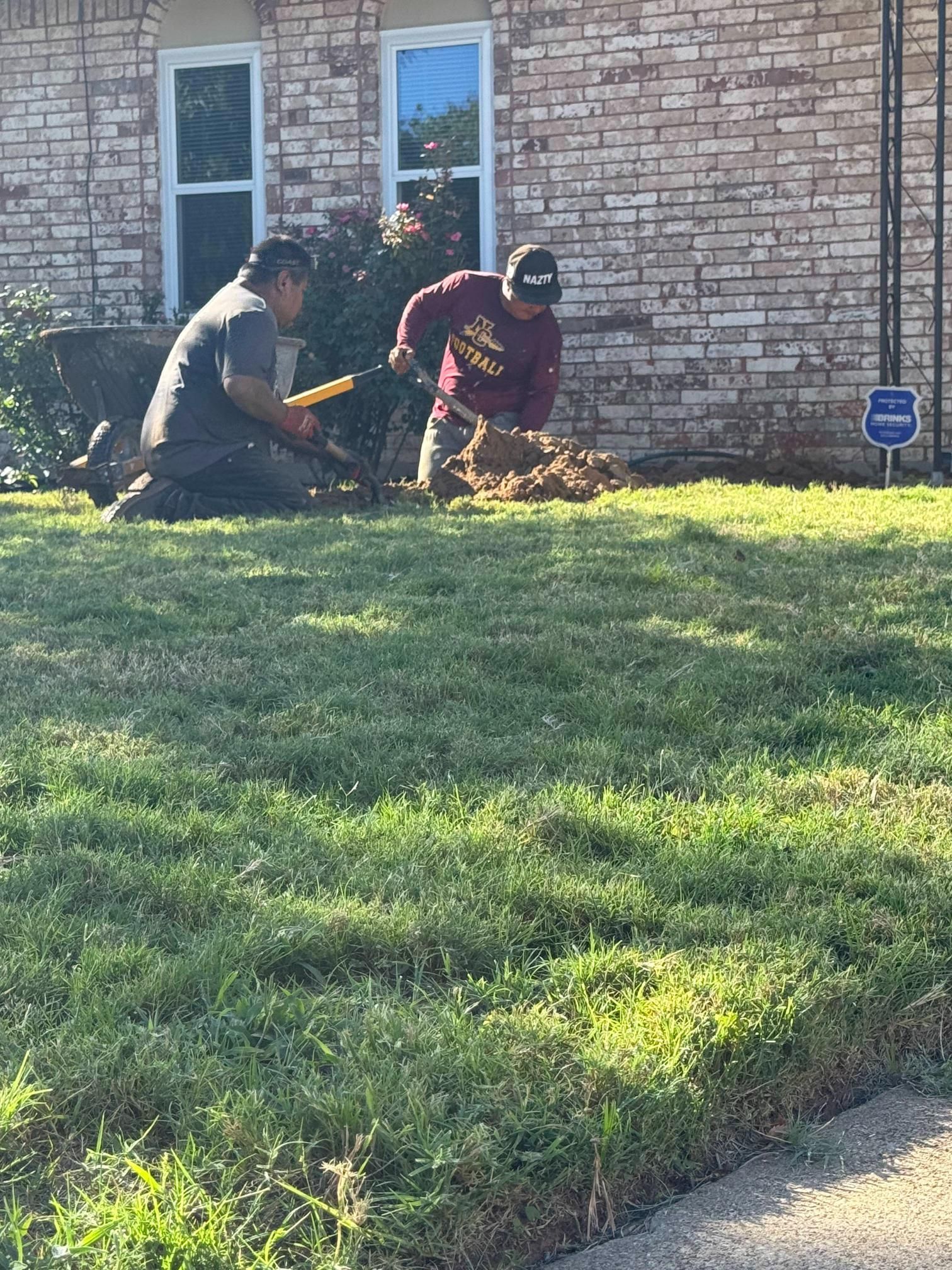 Two people digging in grass near a brick building.