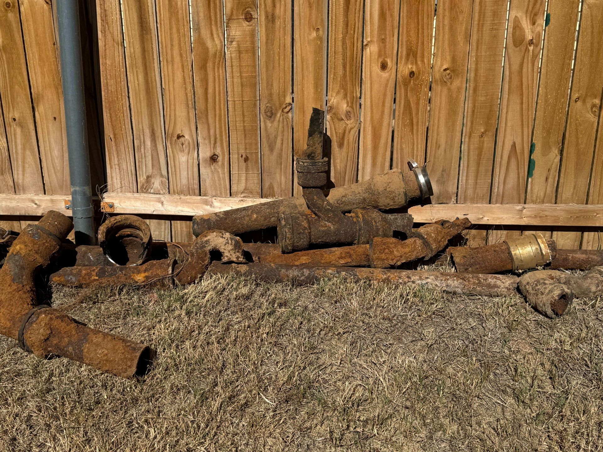 Pile of rusty pipes and fittings on dry grass in front of a wooden fence.