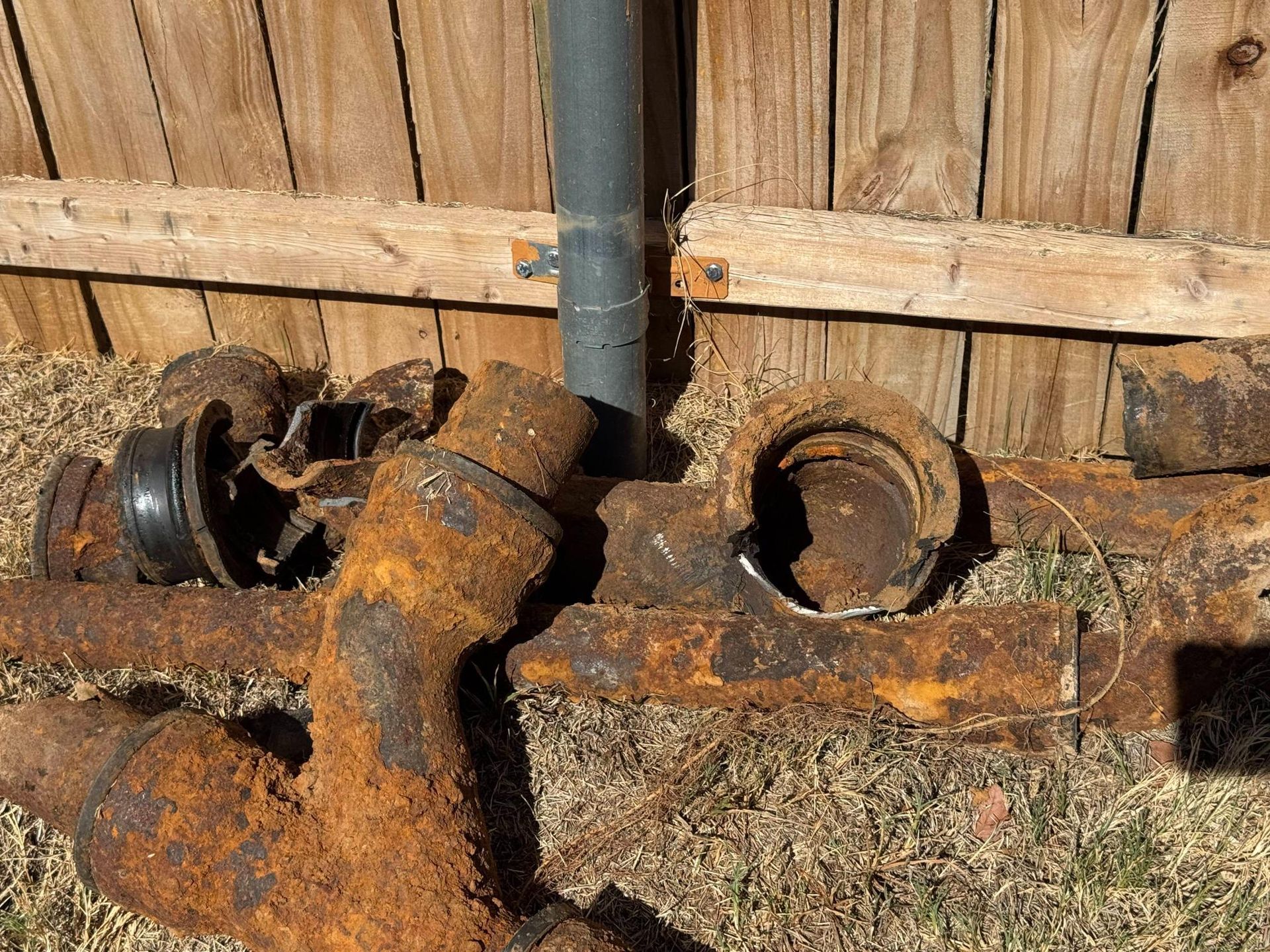 Rusted pipes and fittings lie on grass next to a wooden fence and a dark pipe.